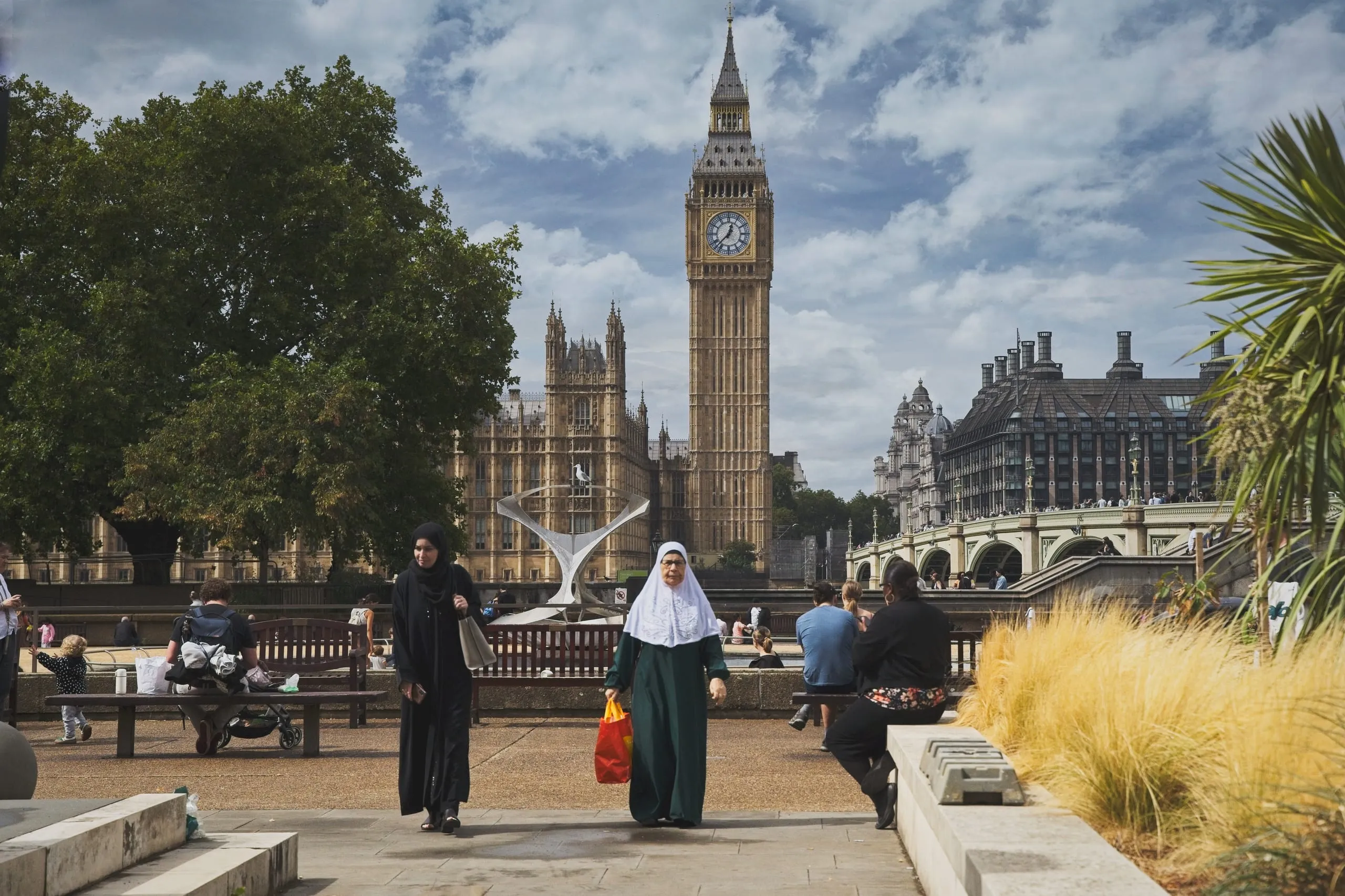 Pedestrians walk through a riverside park in front of Big Ben, the Houses of Parliament, and Westminster Bridge across the River Thames, London, England.