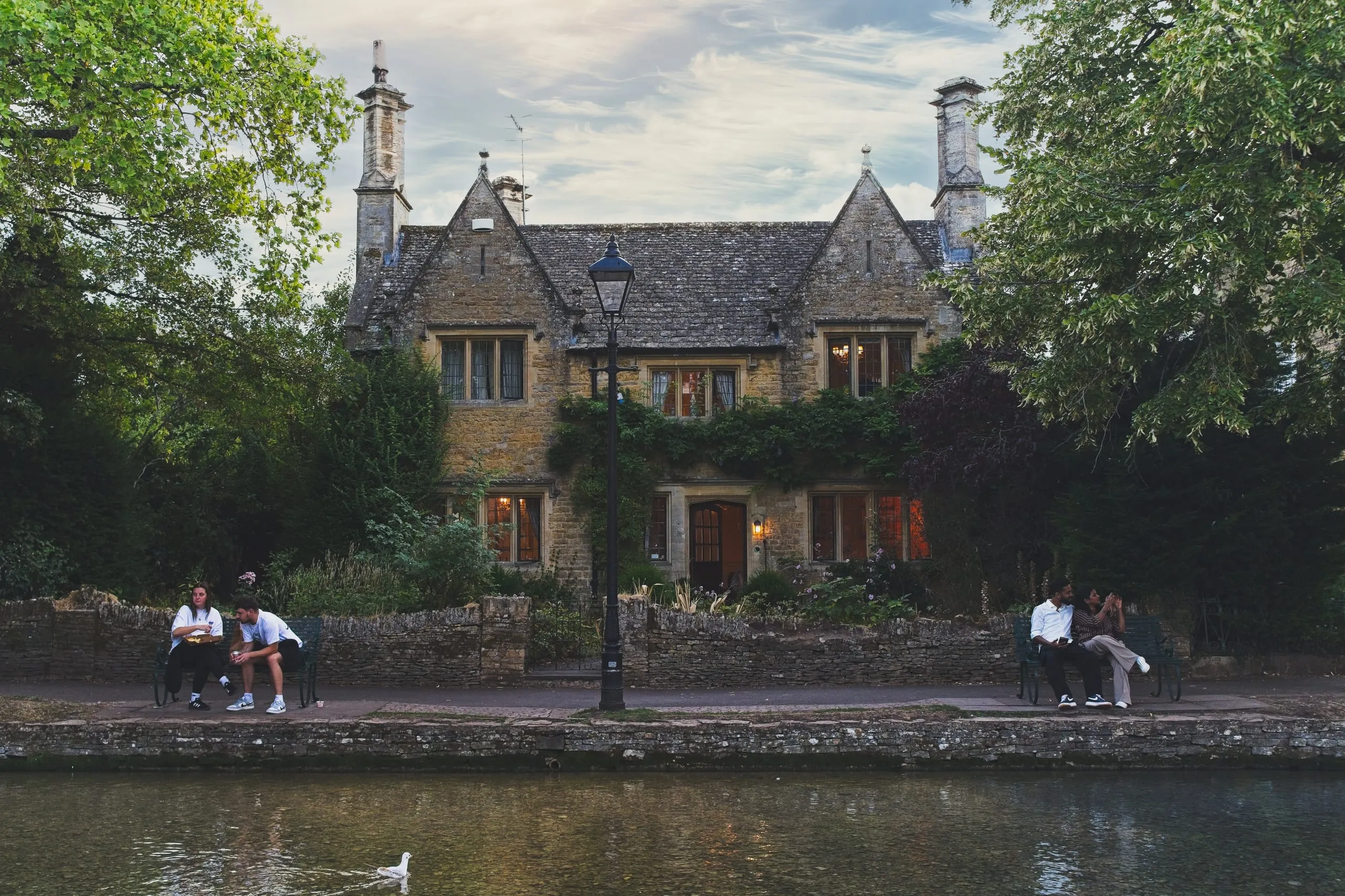 Two couples sit on benches along a riverside path at dusk before a large ivy-covered stone manor house with warmly lit windows, in Bourton-on-the-Water, England.