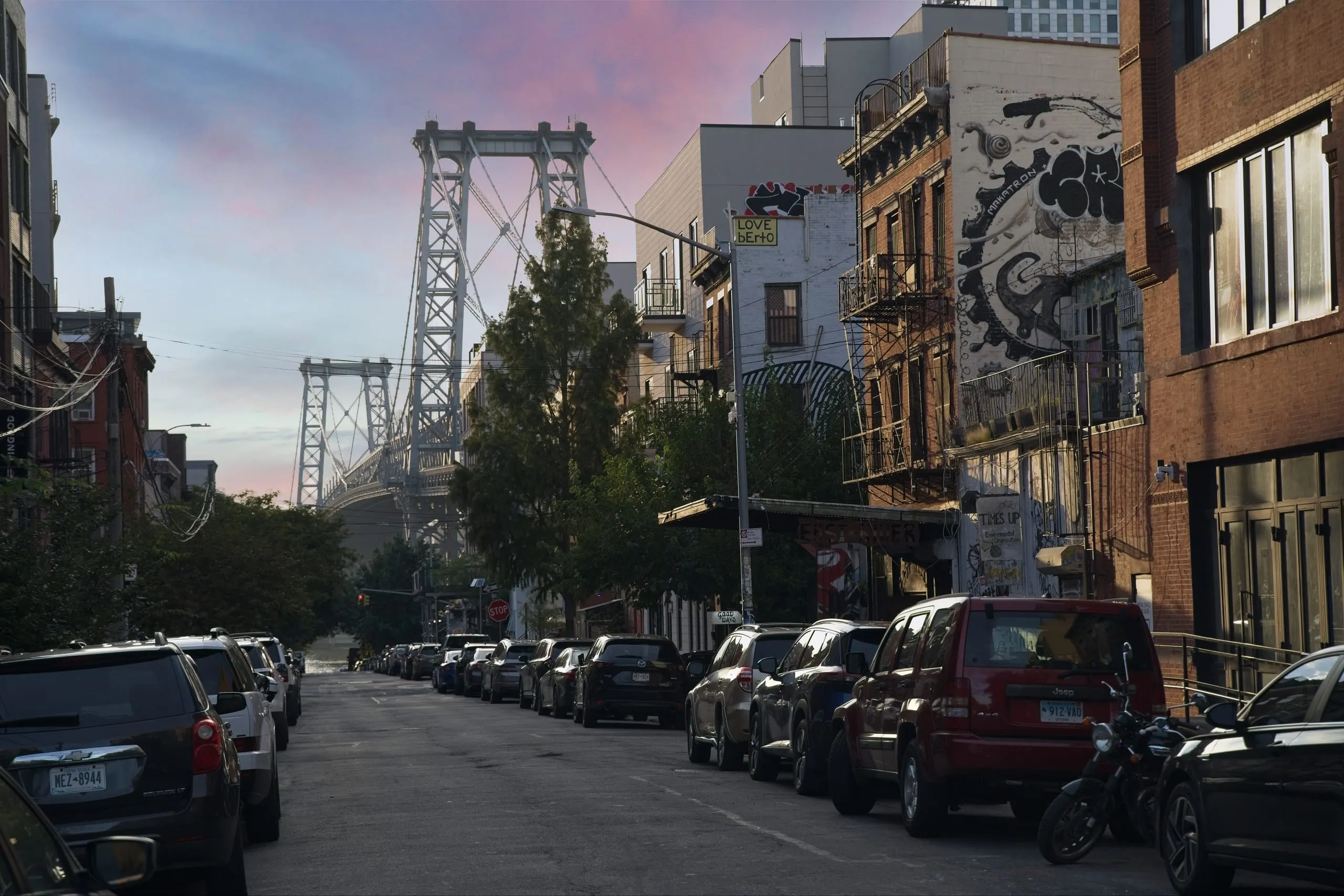 A color photograph of a quiet Williamsburg, Brooklyn street lined with parked cars at dusk, a mural-covered building on the right, and a tower of the Williamsburg Bridge rising against a pink sky in the distance.