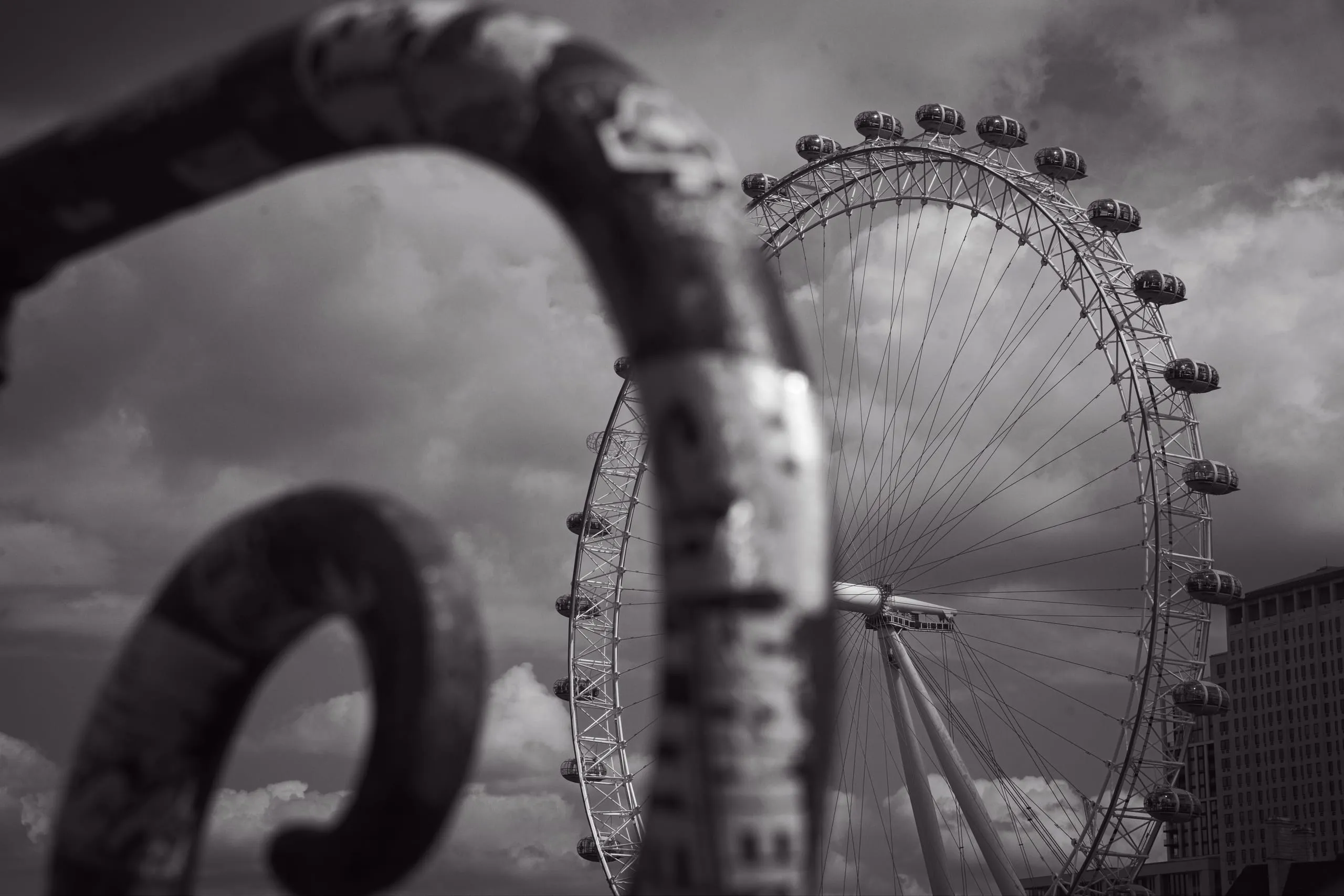 Black and white photograph of the London Eye ferris wheel against a moody sky, seen through the blurred curves of a foreground metal sculpture.