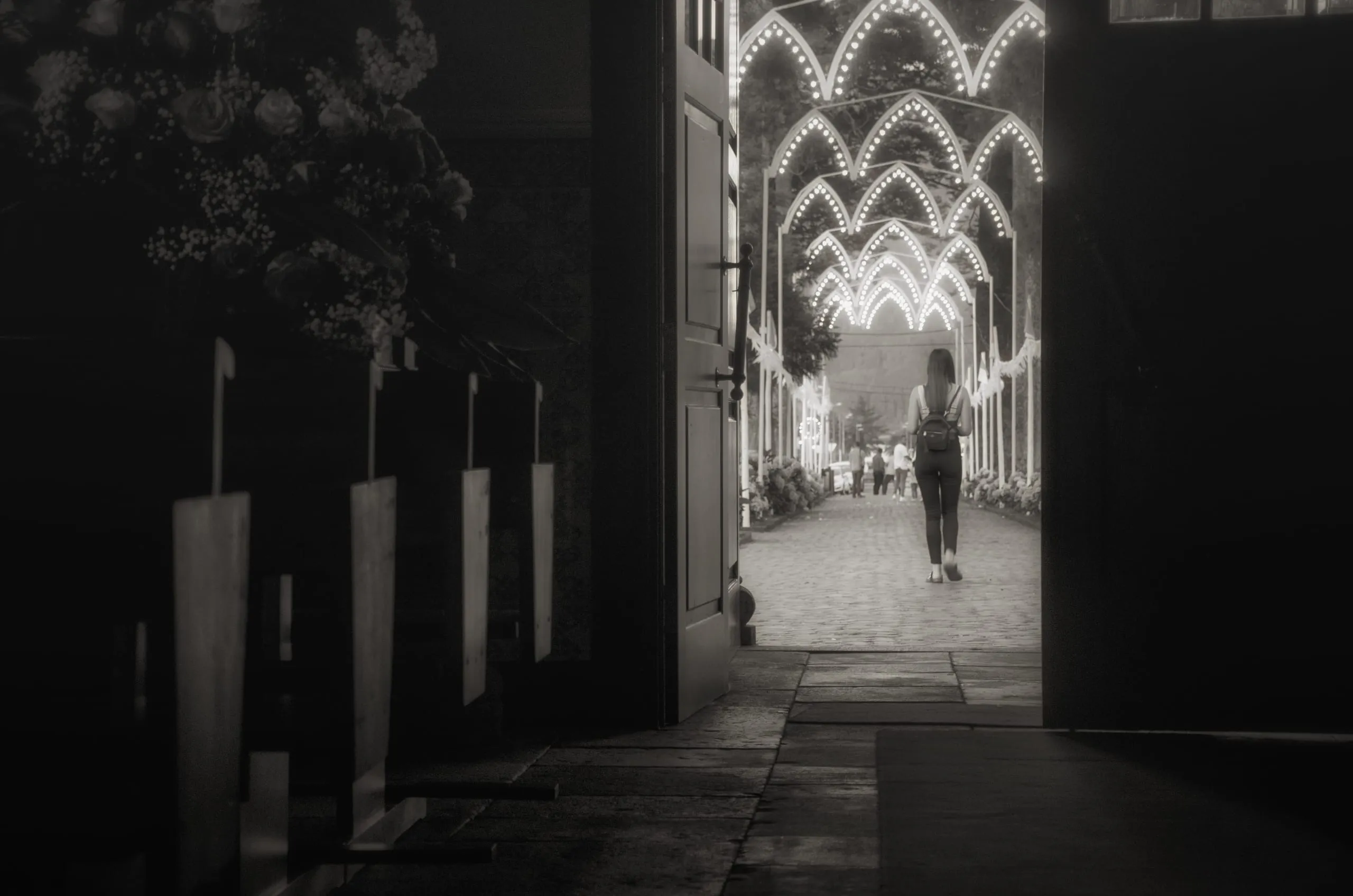 Black-and-white shot from a dark doorway looking out to a woman with a backpack walking a cobbled path beneath rows of lit festival light-arches.