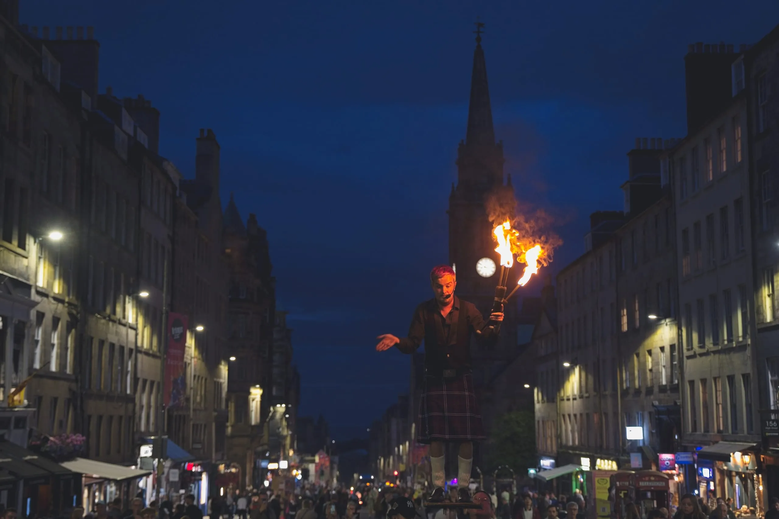 A color photograph of a kilted fire performer balanced on a platform on Edinburgh's Royal Mile at night, torches ablaze in front of a floodlit clock tower.