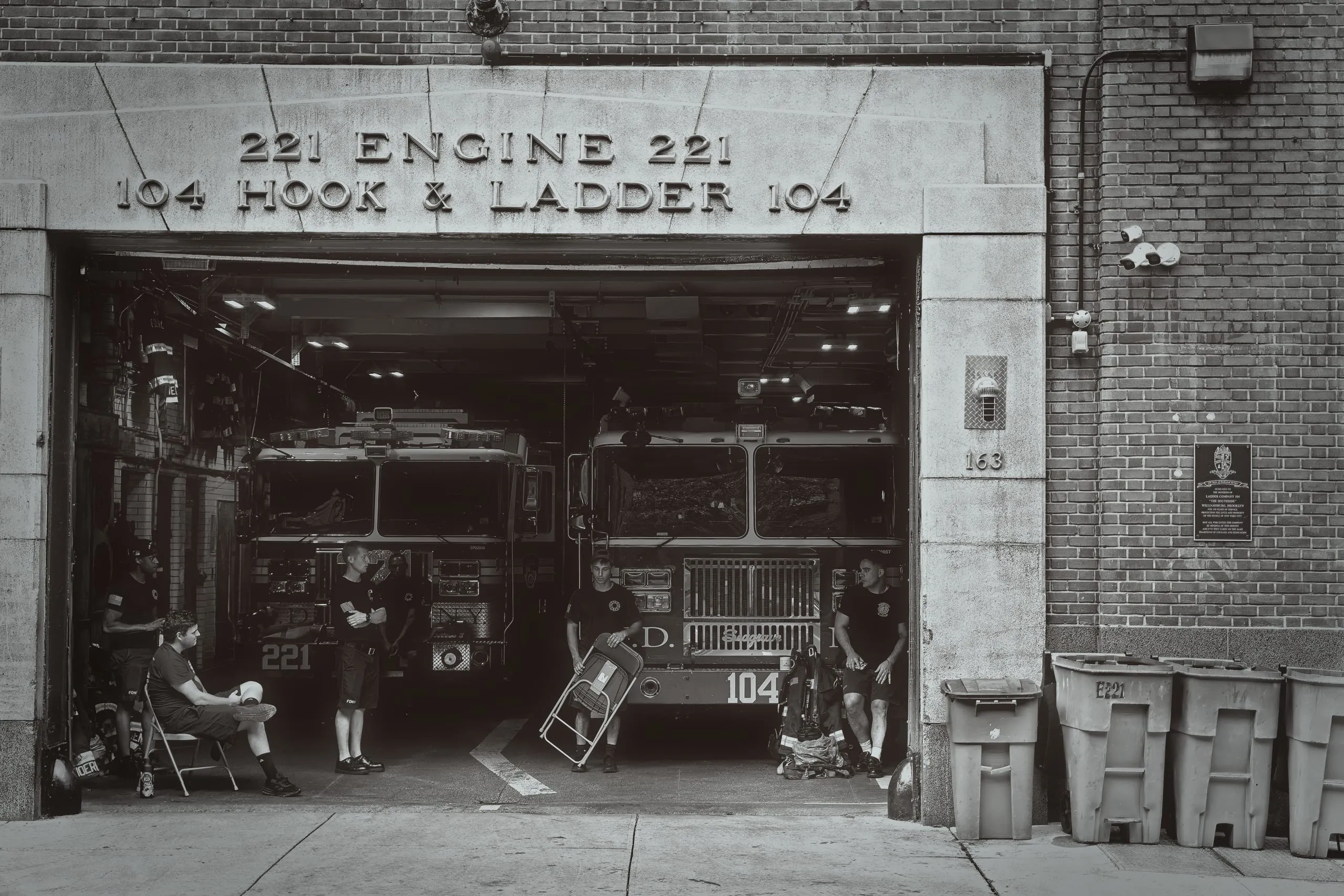 Black and white photograph of firefighters standing beside fire trucks inside the open garage of a Brooklyn firehouse, with carved stone lettering reading Engine 221 and Hook and Ladder 104 above the entrance.