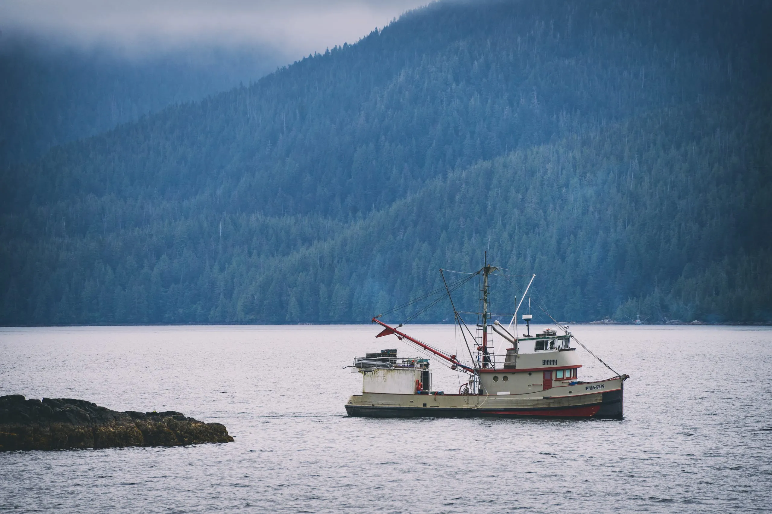 A white and red fishing trawler sits at anchor on calm water below a fog-topped forested mountain slope.