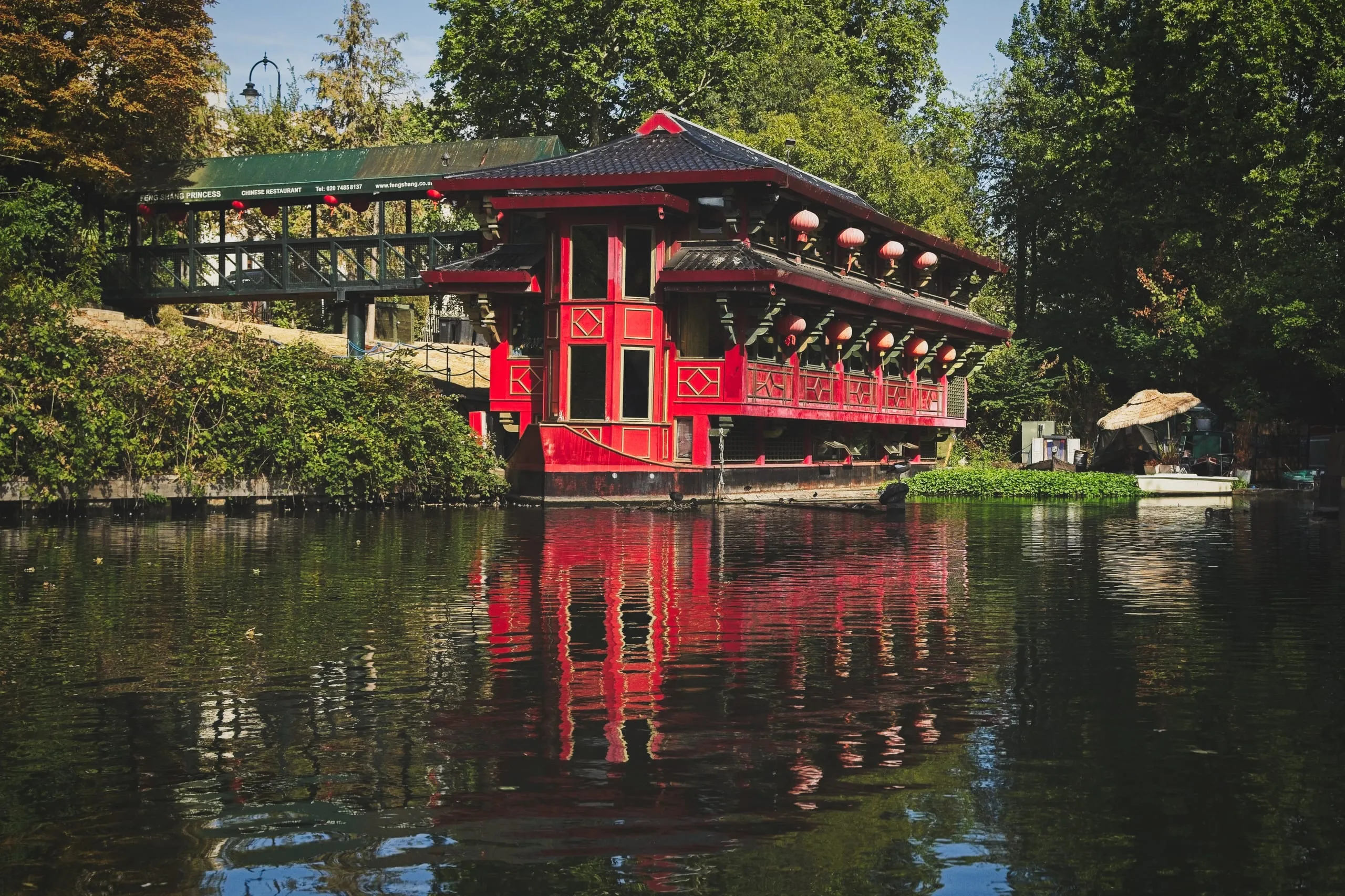 Color photograph of the red pagoda-style Feng Shang Princess floating Chinese restaurant on Regent's Canal in London, surrounded by trees and reflected in the still water.