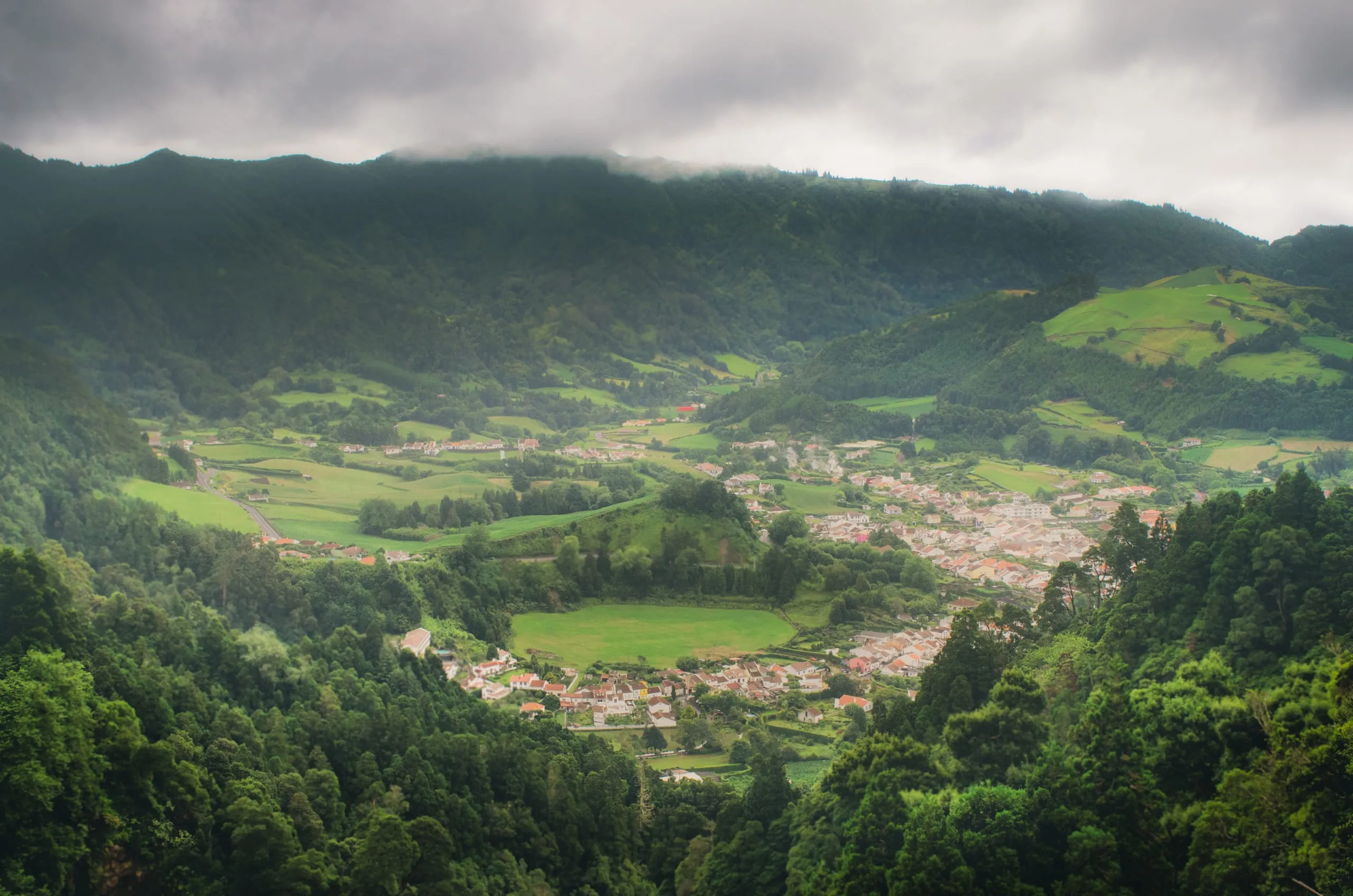 A red-and-white village spreads across a green valley floor beneath cloud-shrouded forested mountains.