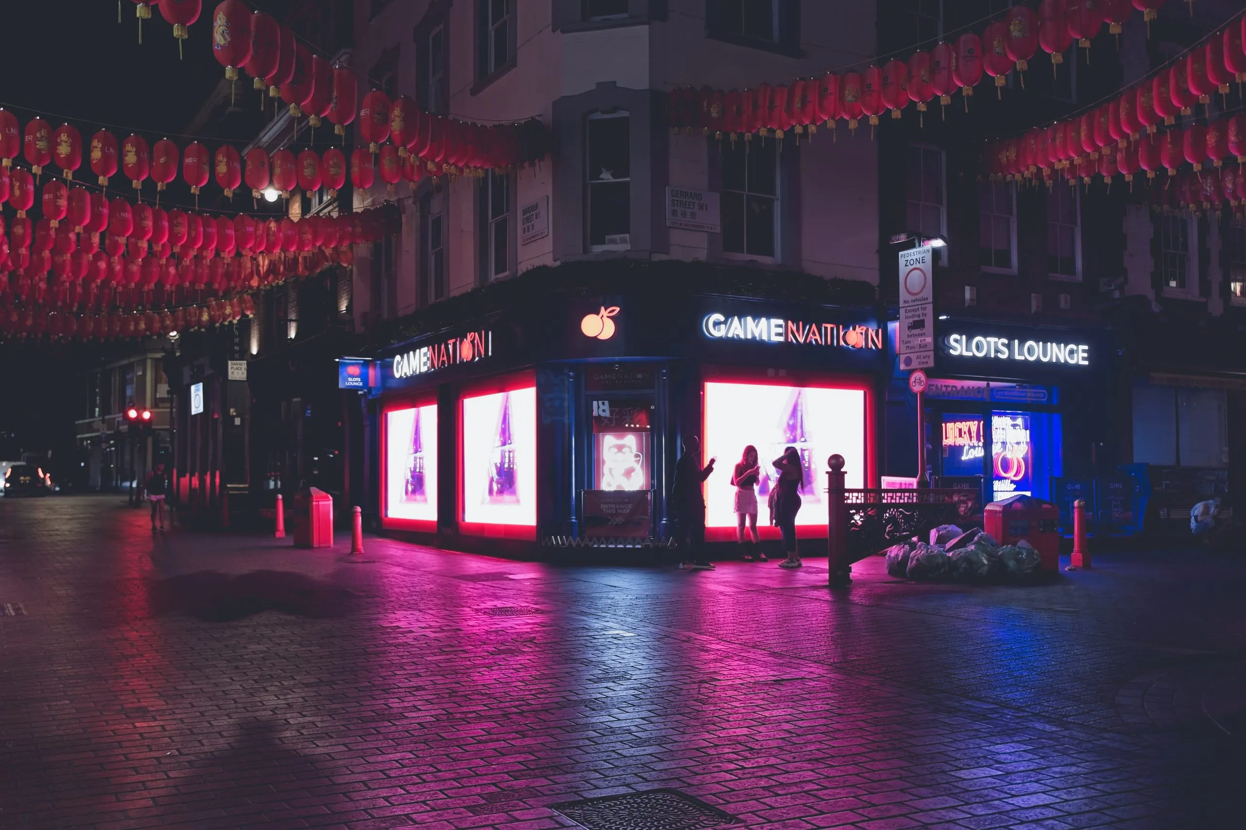 Photograph of a nighttime Chinatown street corner marked Gerrard Street, strung with red lanterns above a glowing pink arcade and slots lounge storefront, London, England.
