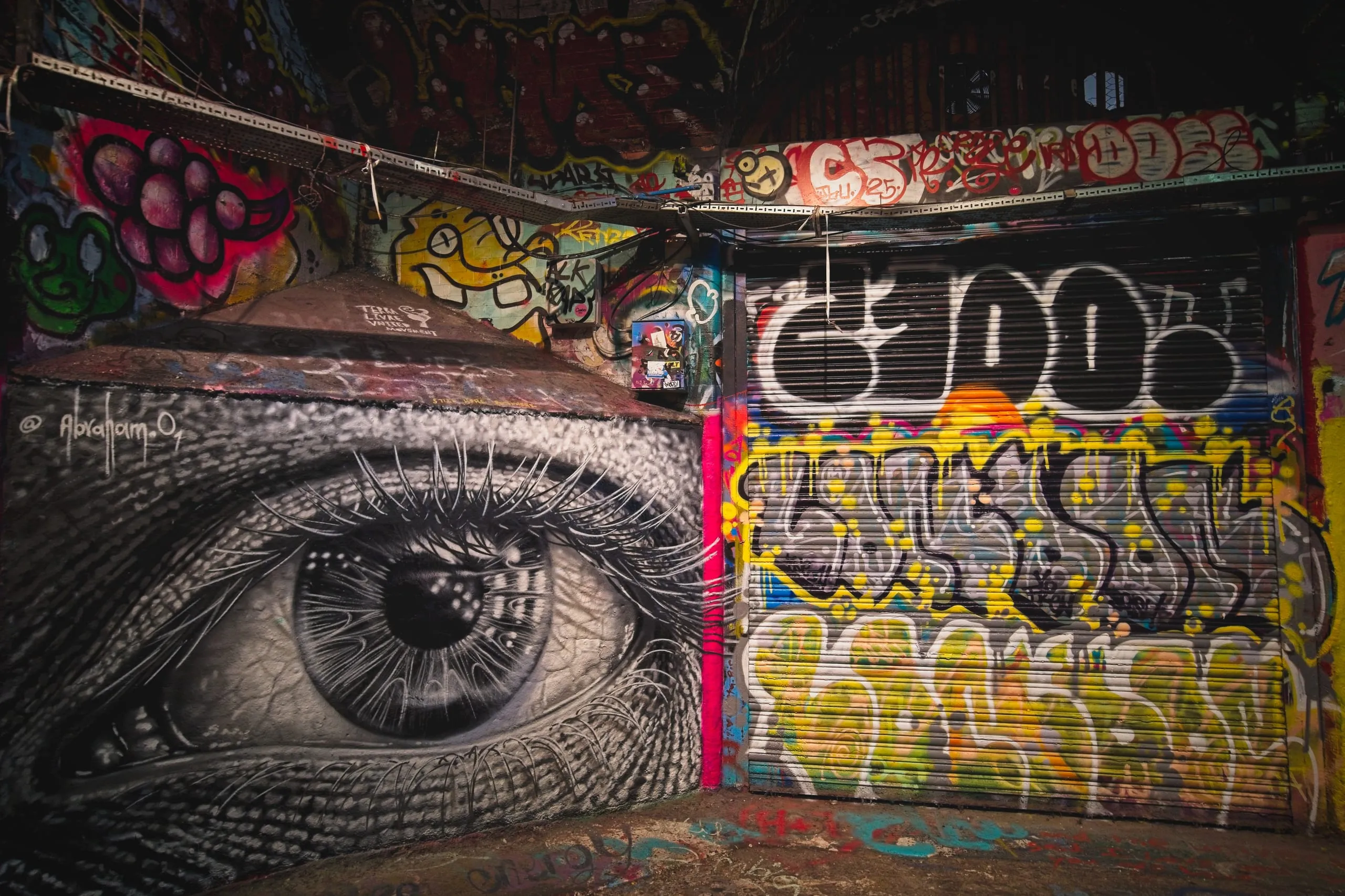 Photograph of a hyperrealistic black-and-white mural of a giant human eye on a wall in London, positioned beside a colorfully graffitied roller shutter door.