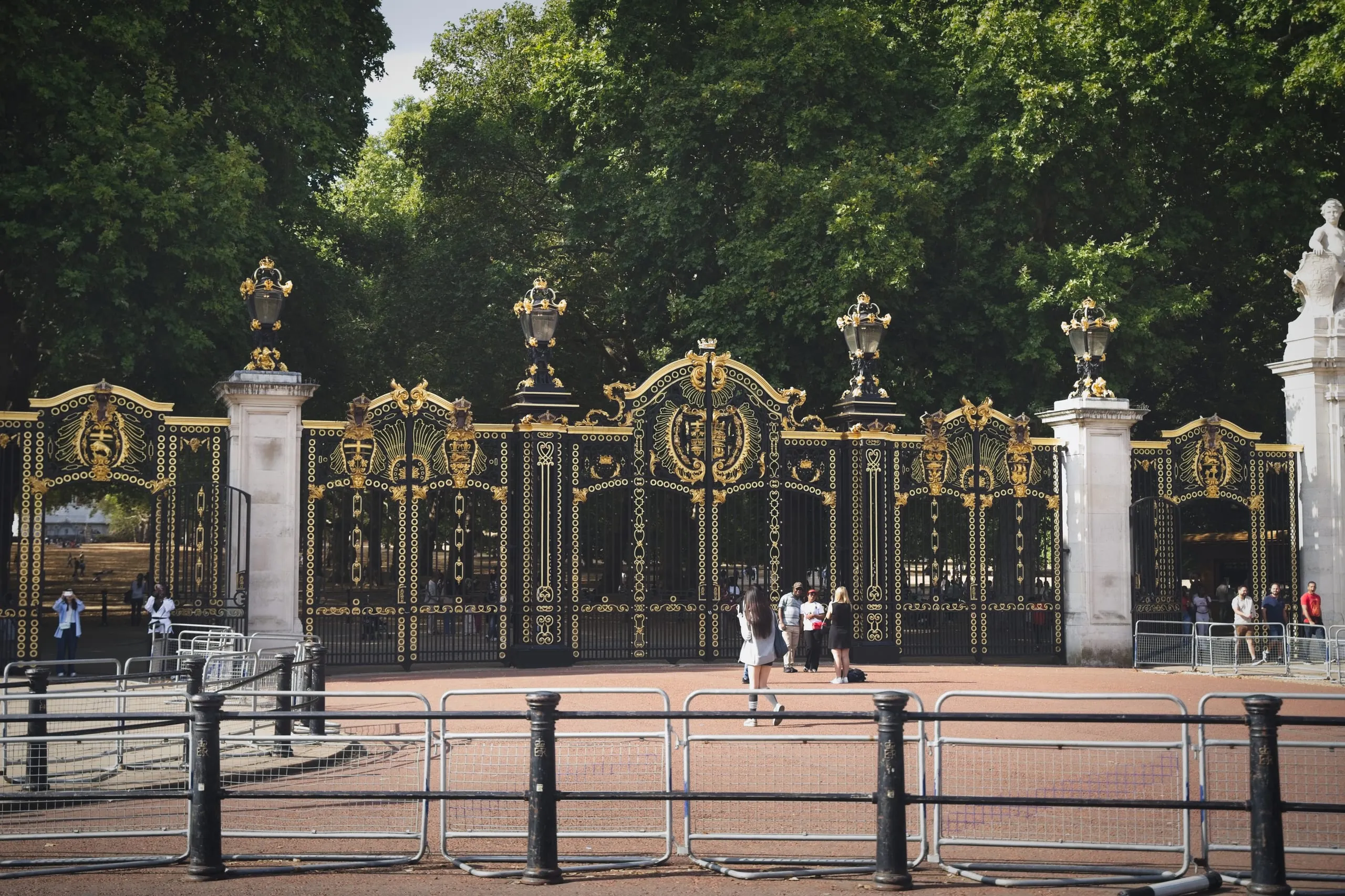 A photograph of the ornate black-and-gold Canada Gate at Green Park near Buckingham Palace in London, with tourists gathered near its entrance on a sunny day.
