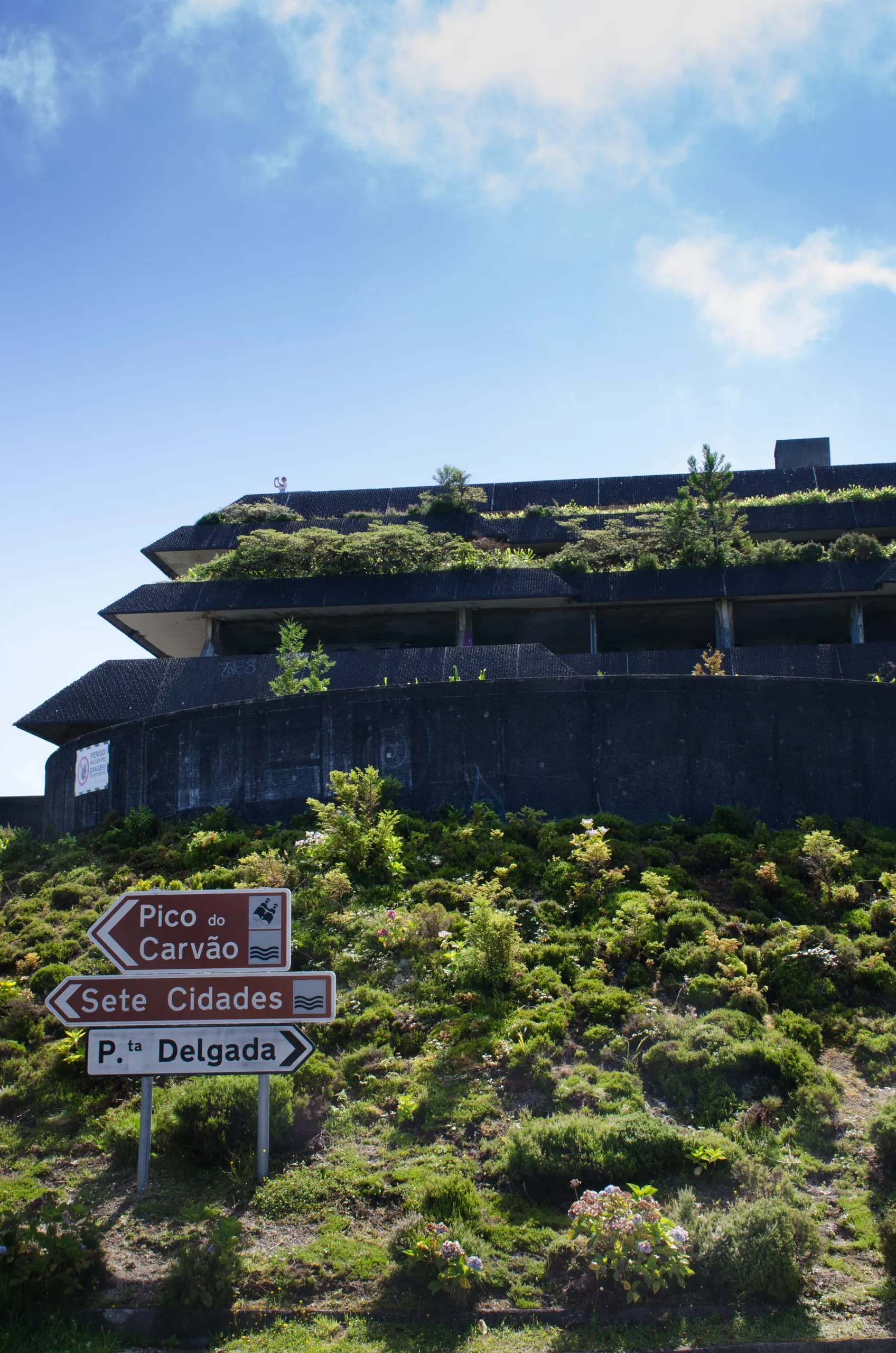 A derelict, overgrown concrete building with plants sprouting from its tiers rises behind a brown directional signpost under a blue sky.