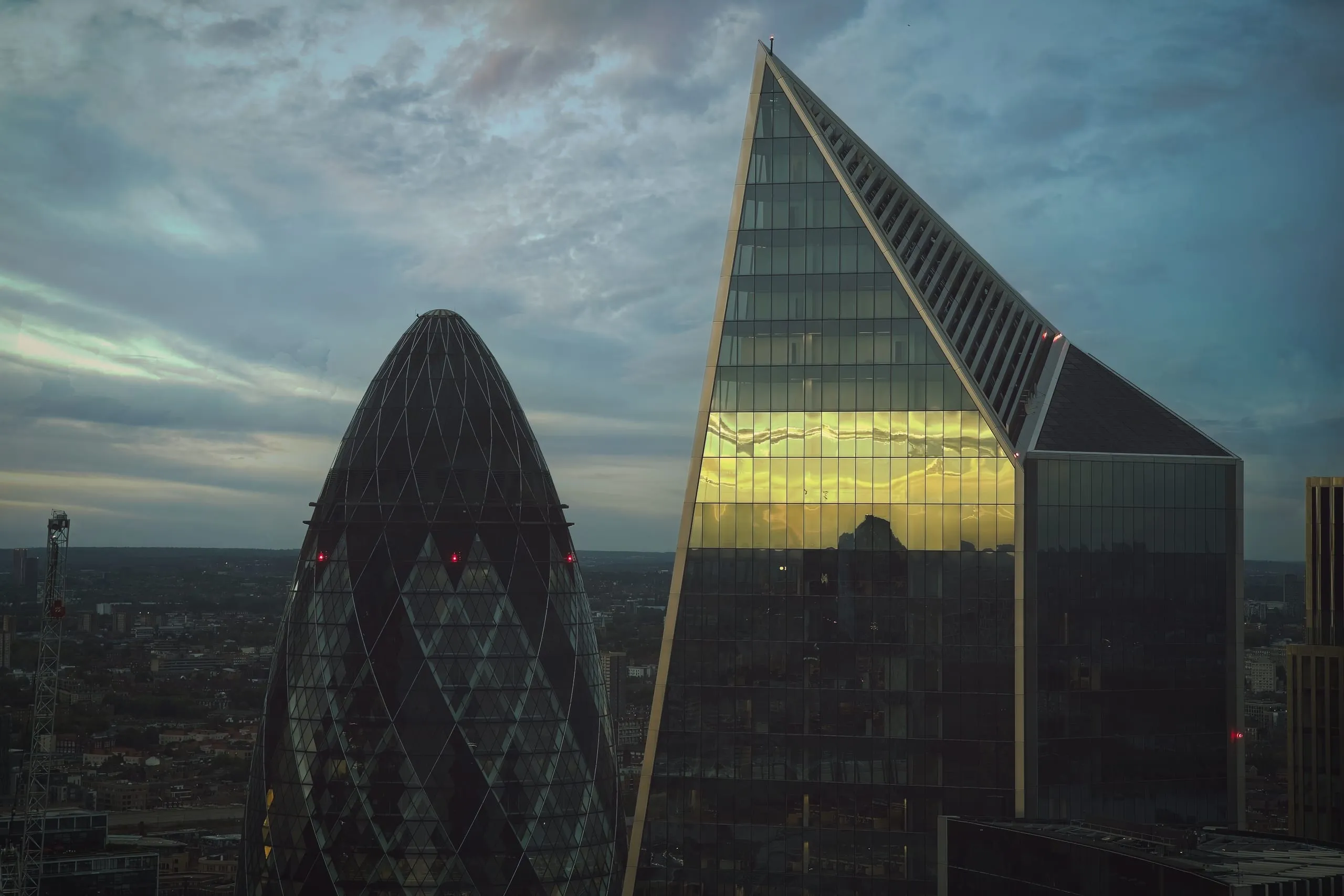A photograph of the Gherkin and the Scalpel skyscrapers in the City of London at dusk, with golden light reflecting off one building's glass facade beneath a cloudy sky.