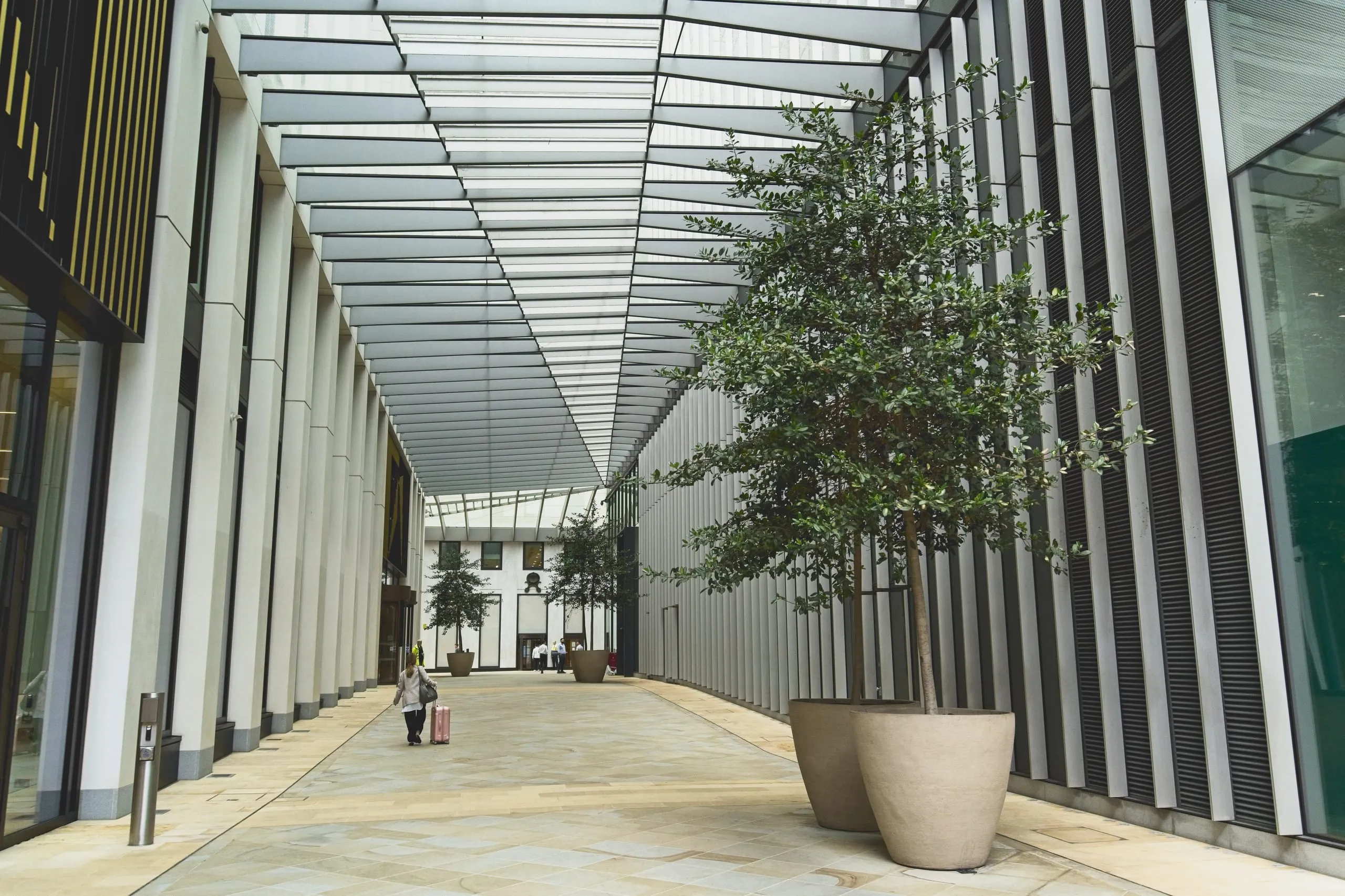 Photograph of a modern glass-roofed pedestrian arcade in London lined with potted trees between two office buildings, a woman pulling a pink suitcase walking down the corridor.