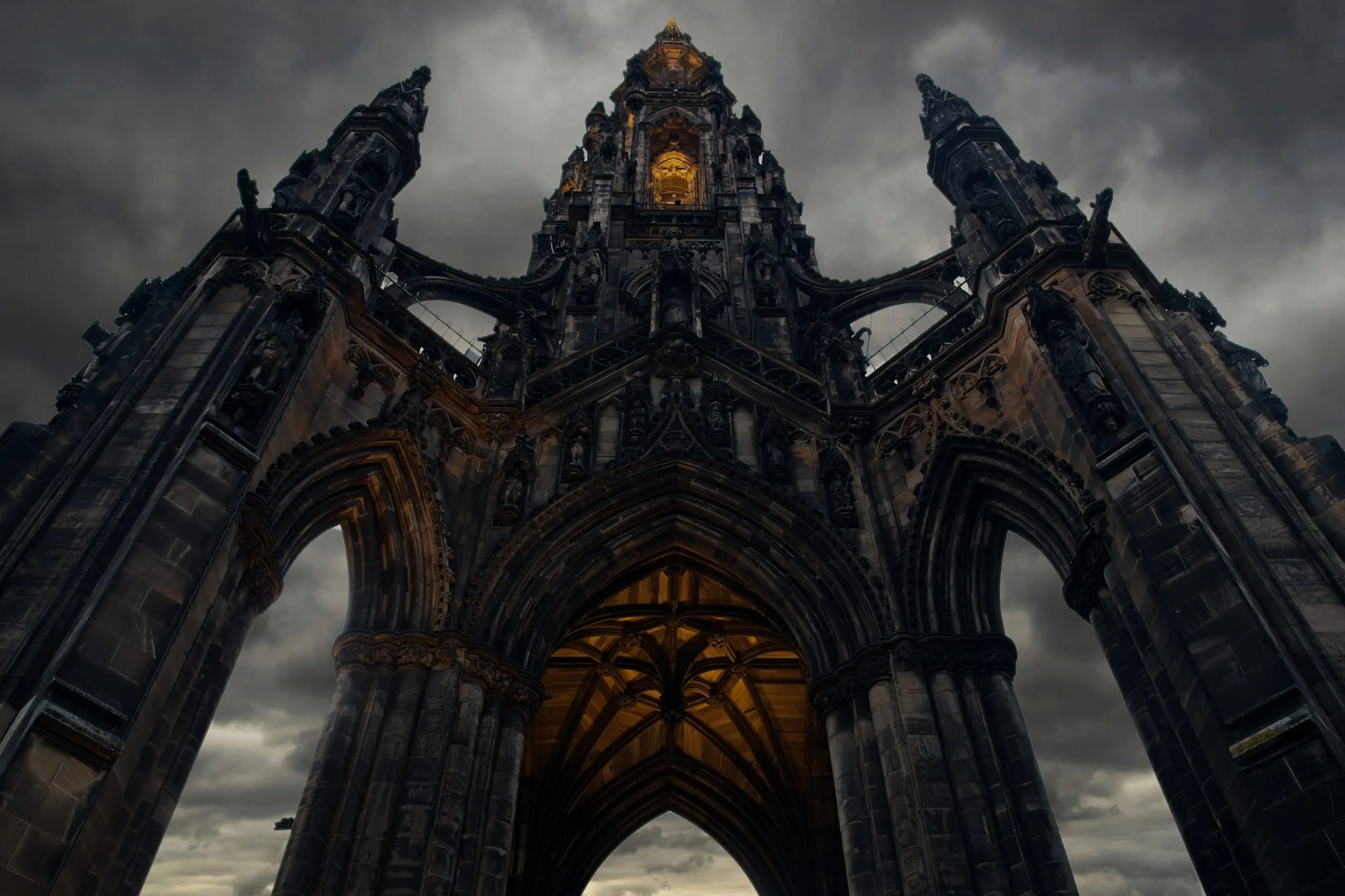 A color photograph looking straight up through the ornate Gothic archways of the Scott Monument in Edinburgh, its interior lit gold against a brooding, stormy sky.