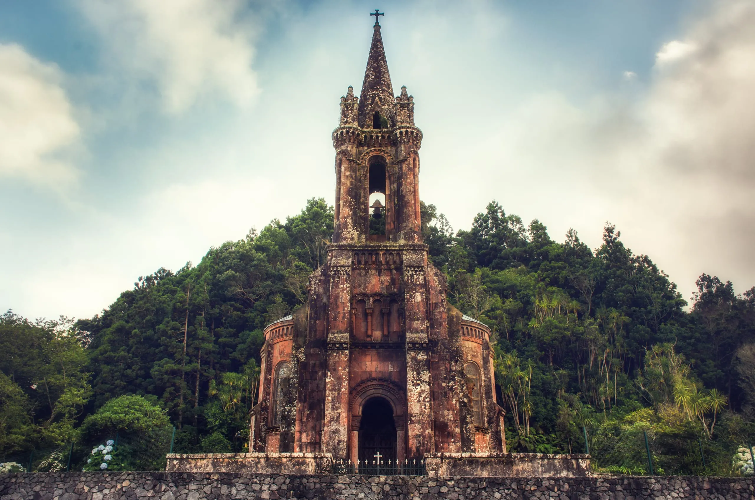 A lichen-stained neo-Gothic stone chapel with a tall pinnacled spire and bell, backed by dense green forest under a cloudy sky.
