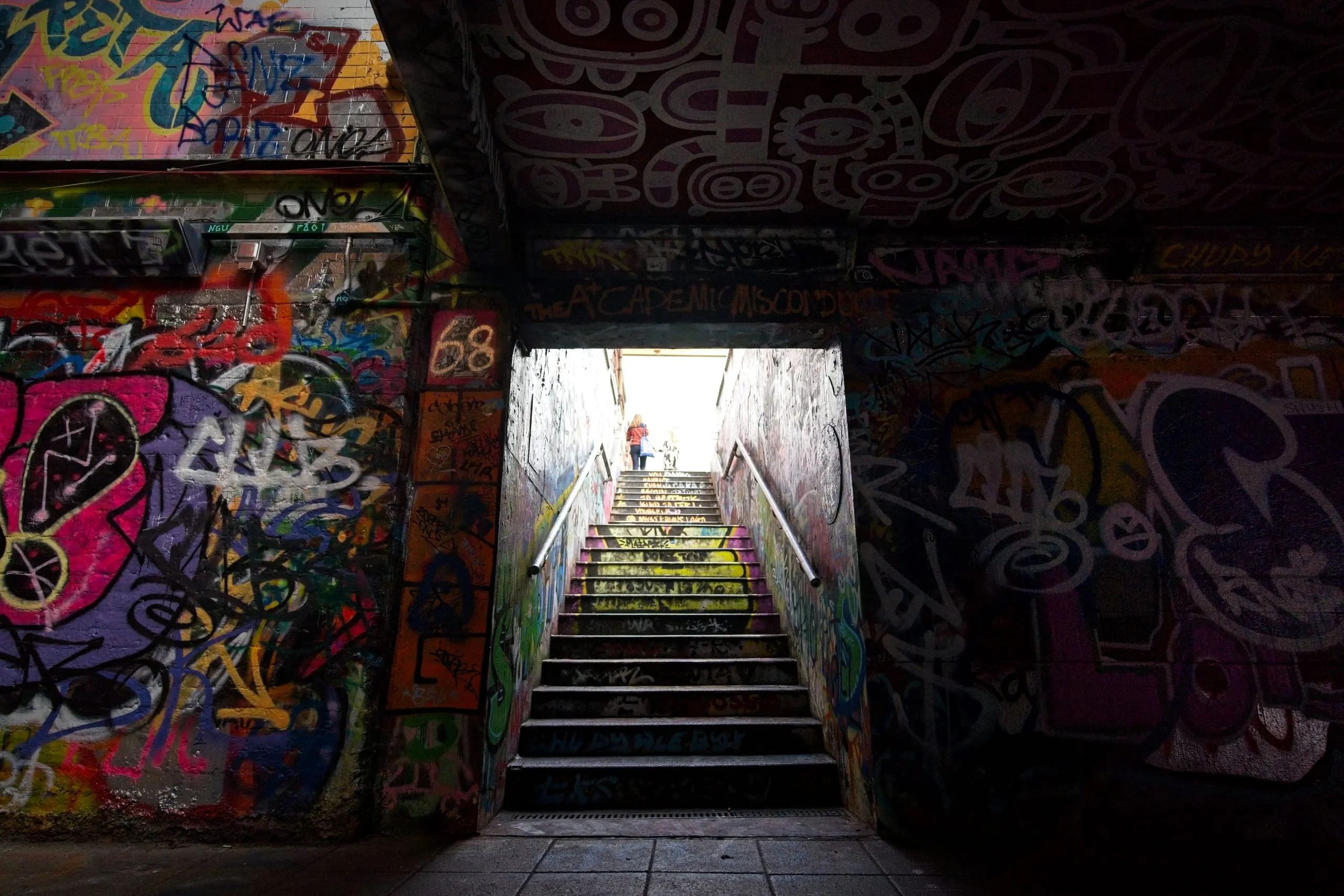 Photograph of a graffiti-covered stairwell in London, dark at the bottom with densely tagged walls, brightening toward a sunlit exit at the top where a woman is climbing the stairs.