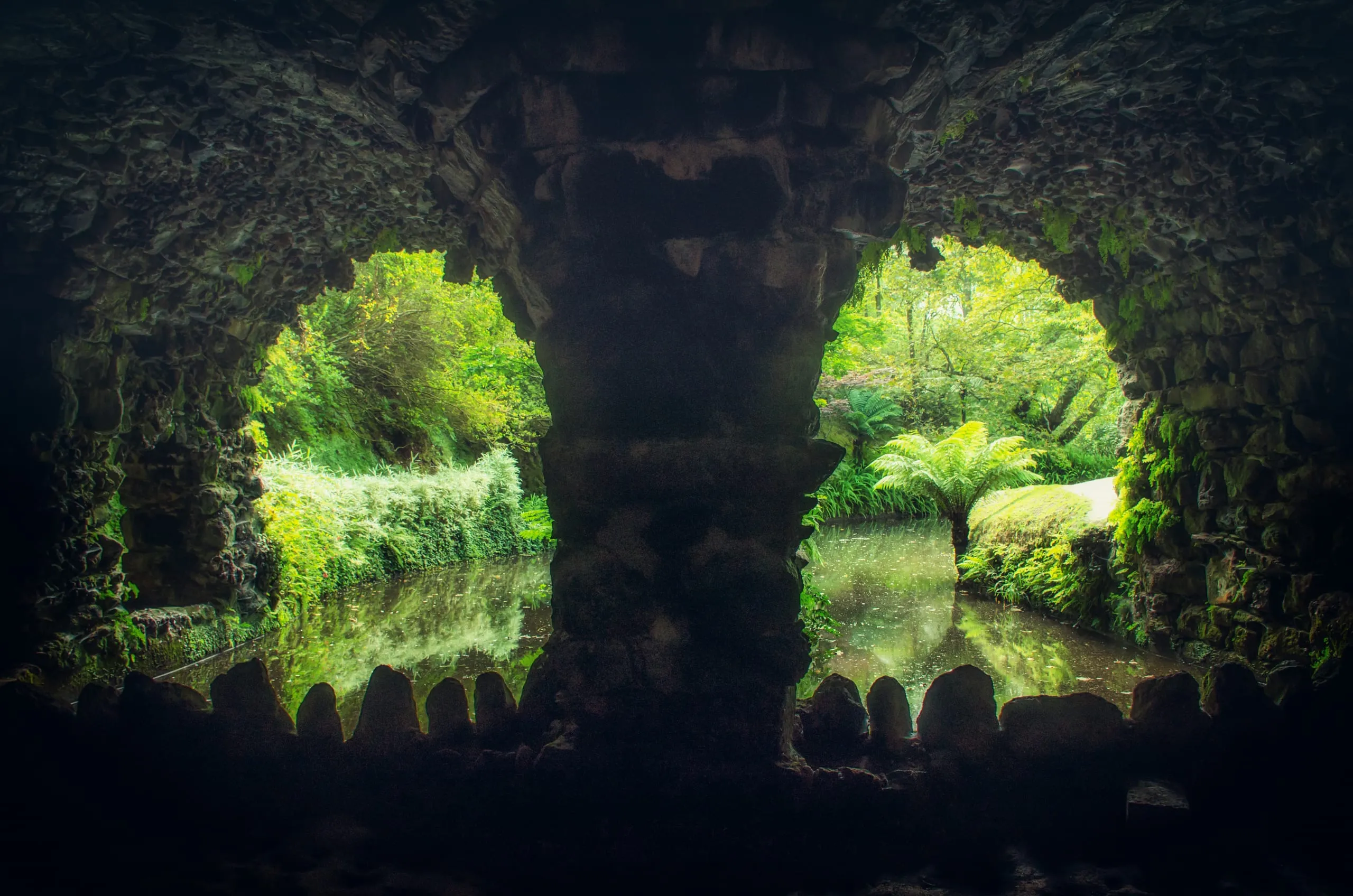 View from inside a dark mossy stone grotto through arched openings onto a still green pond and lush ferns.