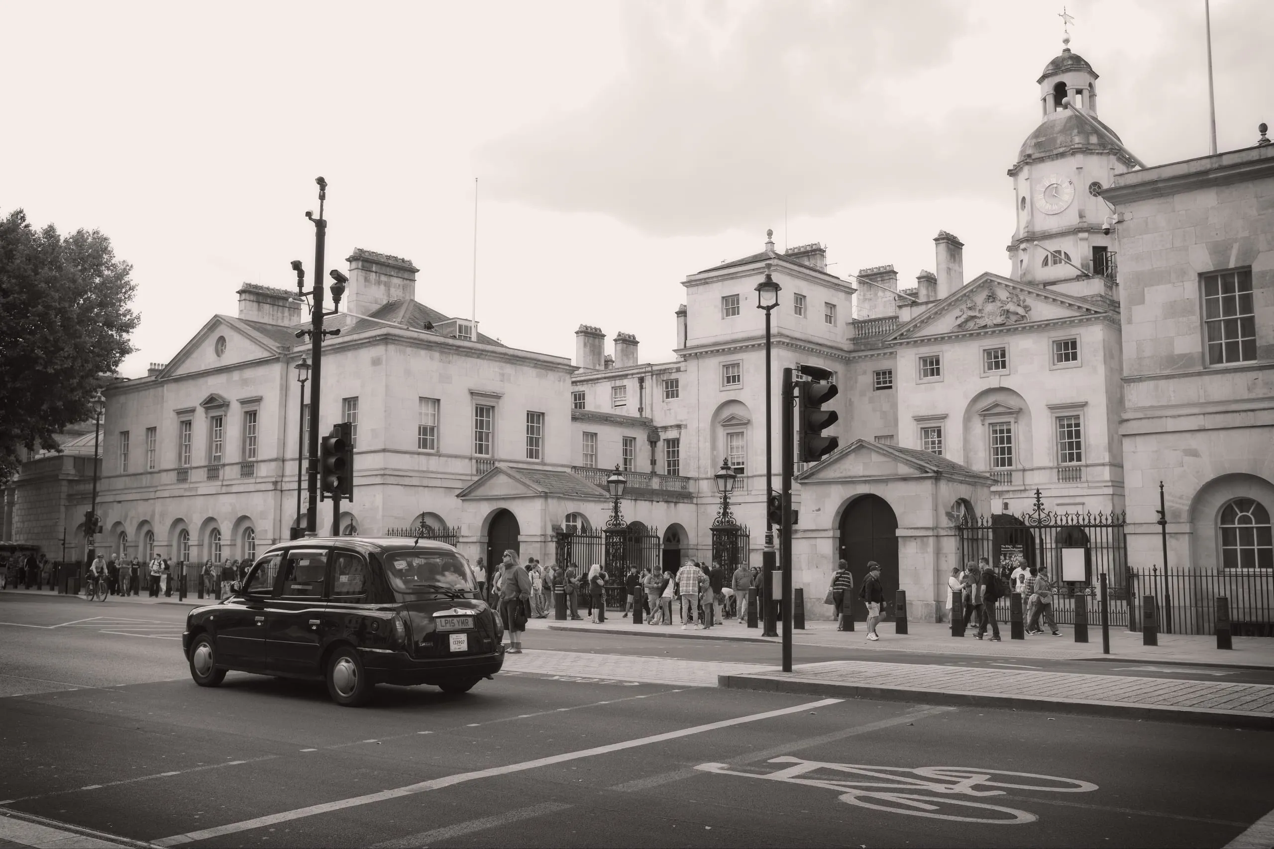 Sepia-toned monochrome photograph of a black London taxi cab passing the Horse Guards building on Whitehall, London, with tourists gathered near the gate beneath a clock tower.