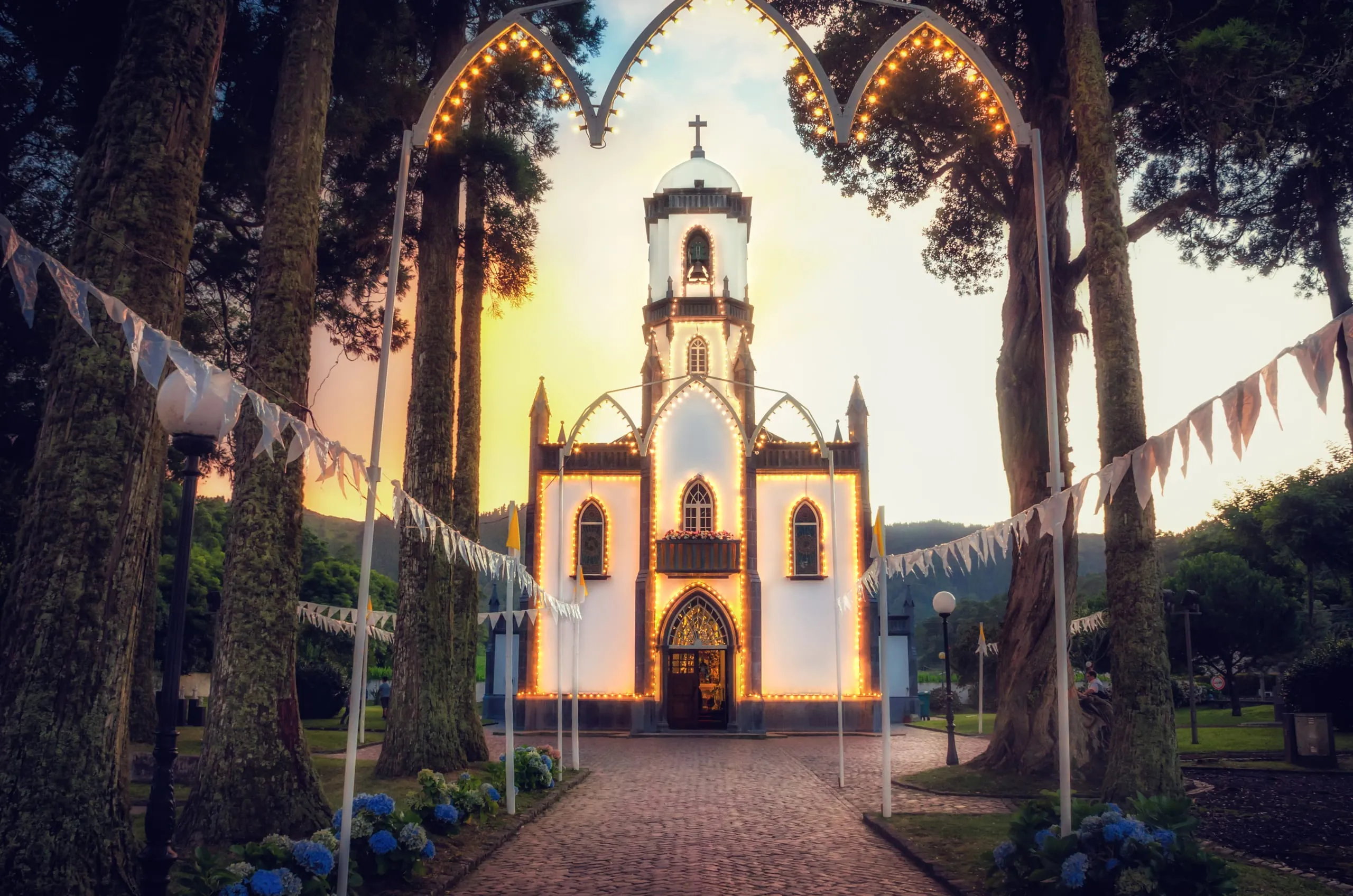 A white gothic chapel outlined in warm festival lights at dusk, framed by tall trees, a lit archway, and bunting over a cobbled path lined with hydrangeas.
