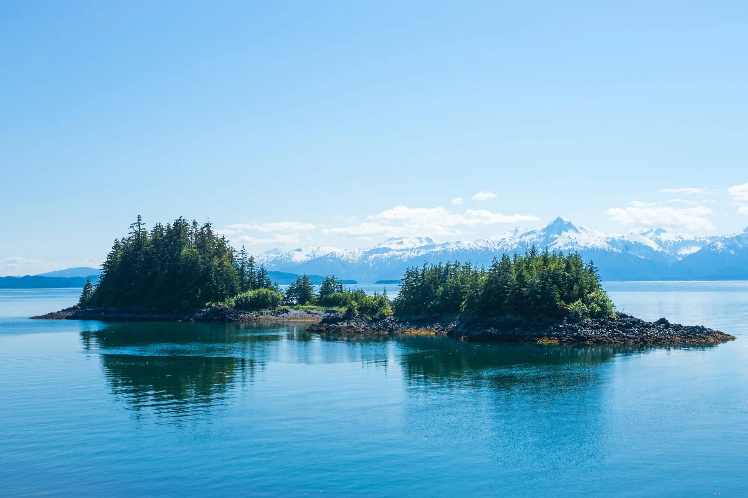 Two small spruce-topped rocky islands reflect in glassy blue water, with a snow-covered mountain range on the far horizon under clear sky.