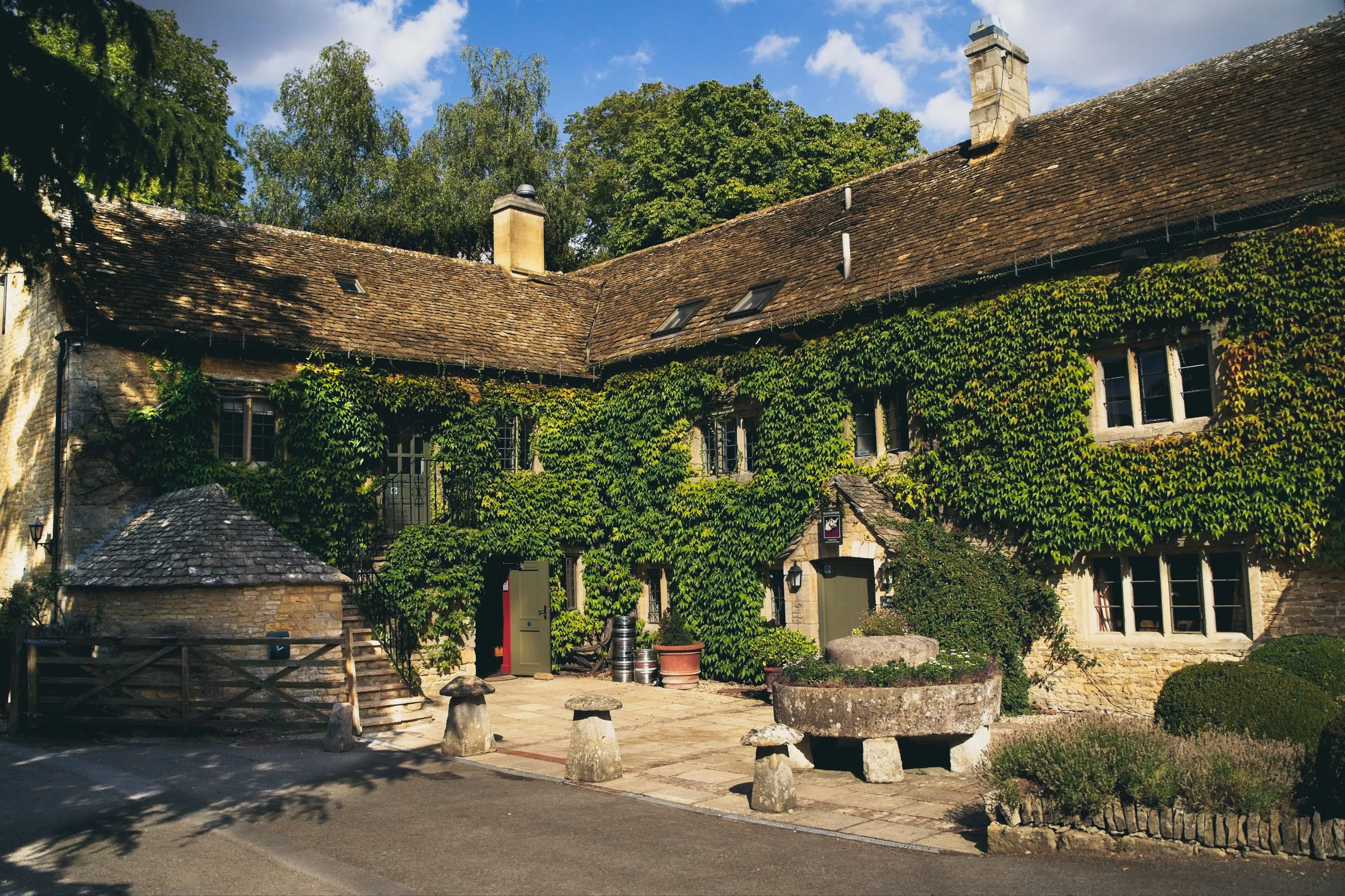 A photograph of an ivy-covered stone building in the Cotswolds, England, with a tiled roof, stone chimneys, and mushroom-shaped staddle stones in the courtyard.