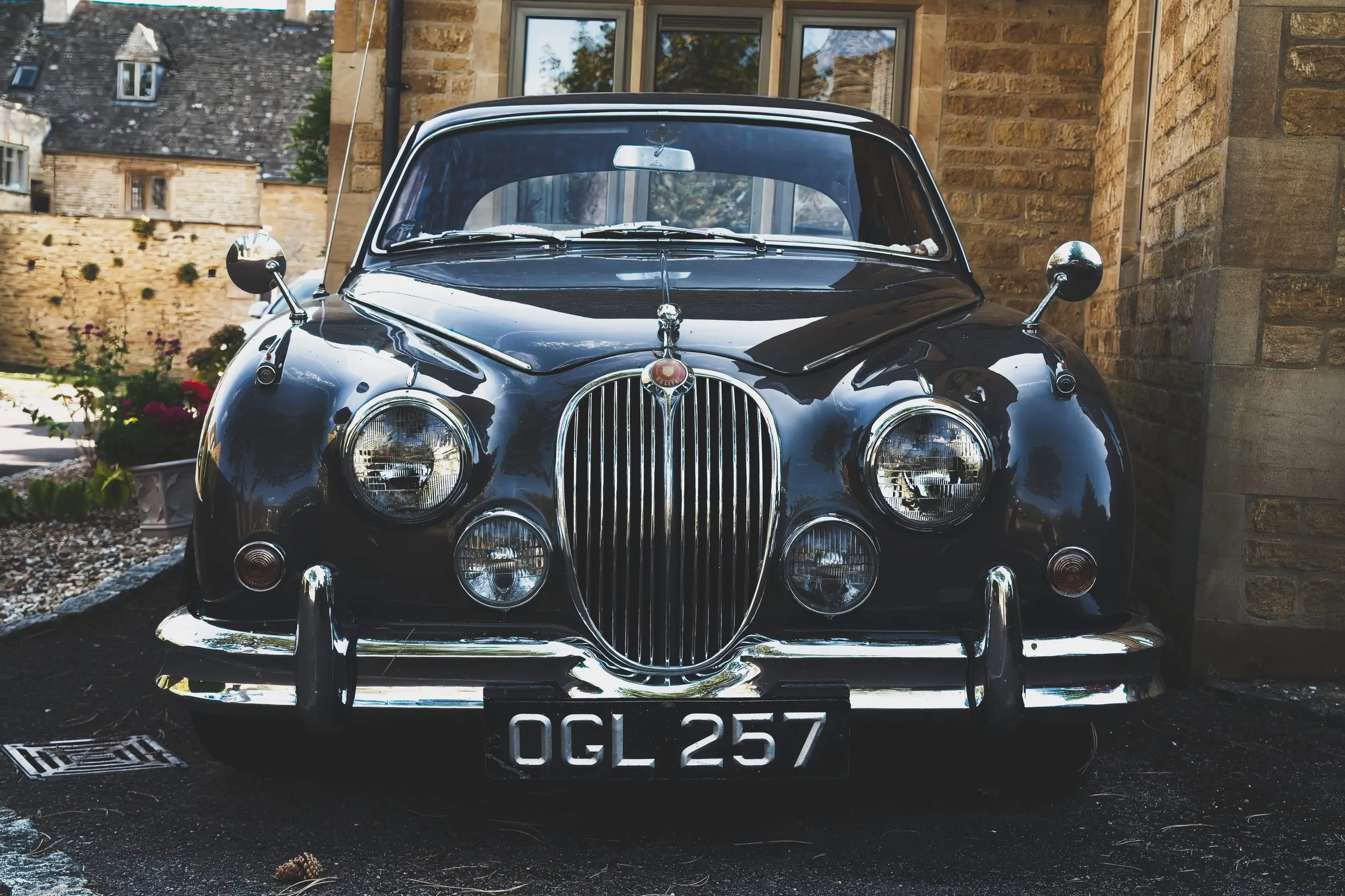 A photograph of the front grille and headlights of a vintage black Jaguar saloon parked in the Cotswolds, England, its license plate reading OGL 257.