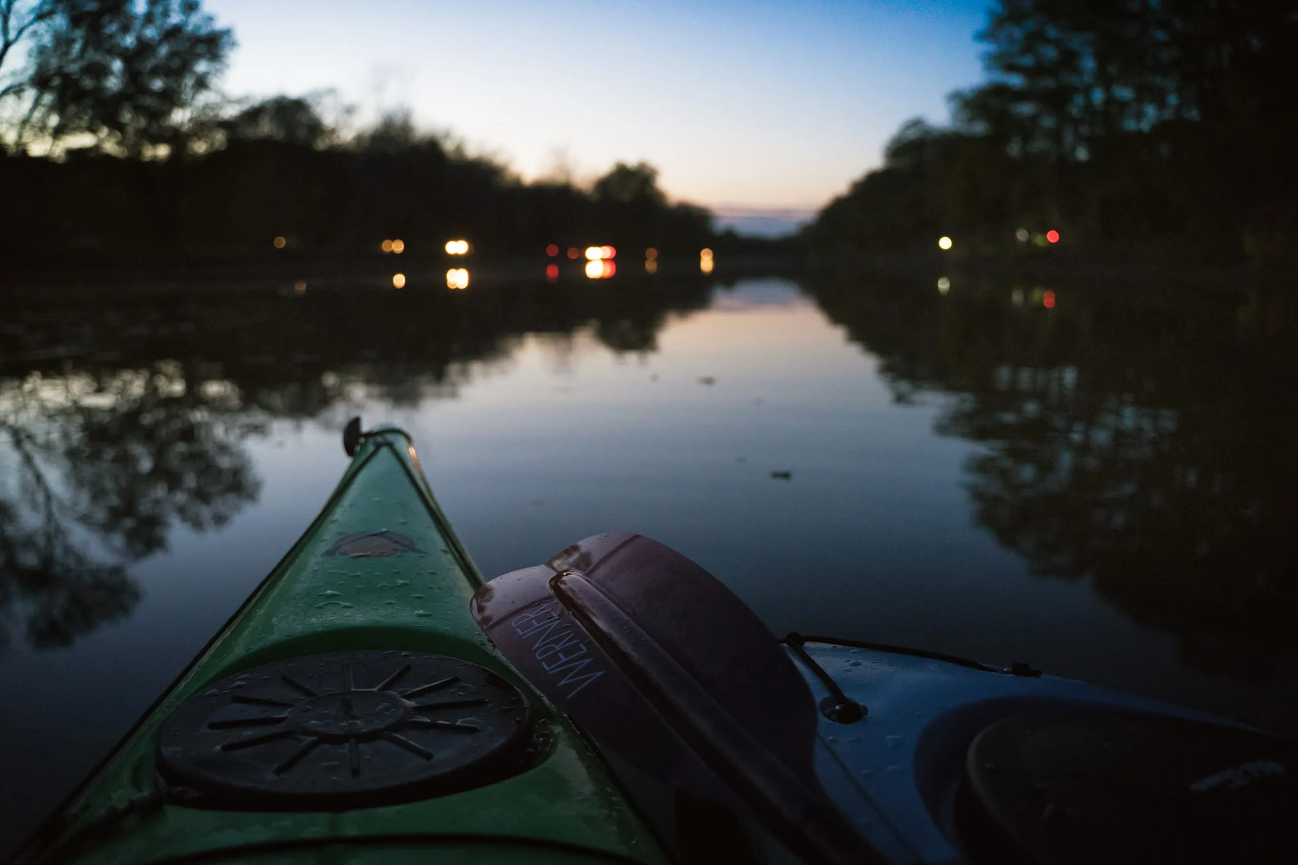 View over a kayak bow at twilight with shoreline lights reflecting on the Erie Canal