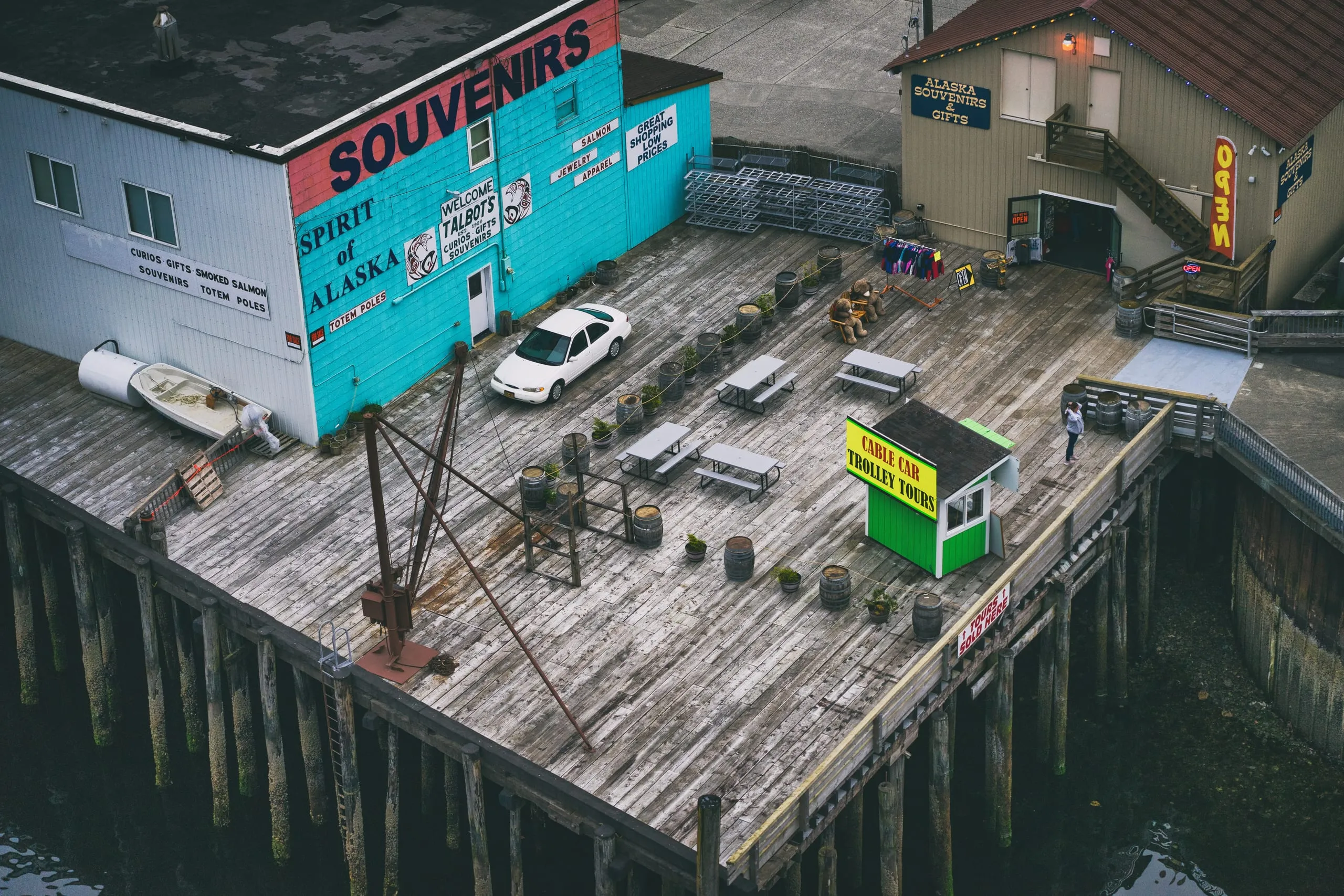 An elevated view of a weathered wooden pier lined with brightly painted souvenir shops, picnic tables, whiskey barrels, and a green ticket booth.