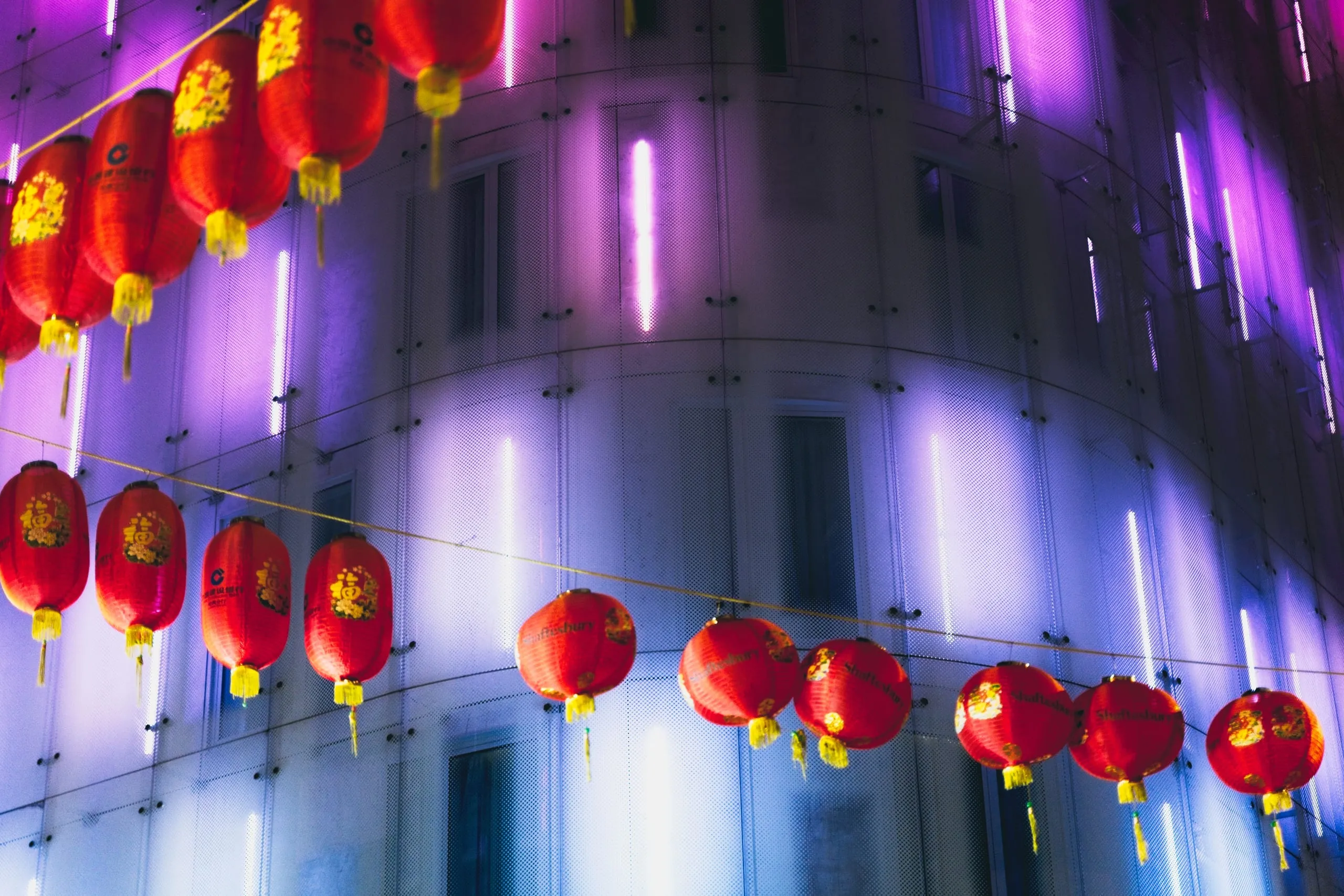 Photograph of strings of red Chinese lanterns hanging against a curved building lit purple and white in London's Chinatown near Shaftesbury Avenue, London, England.