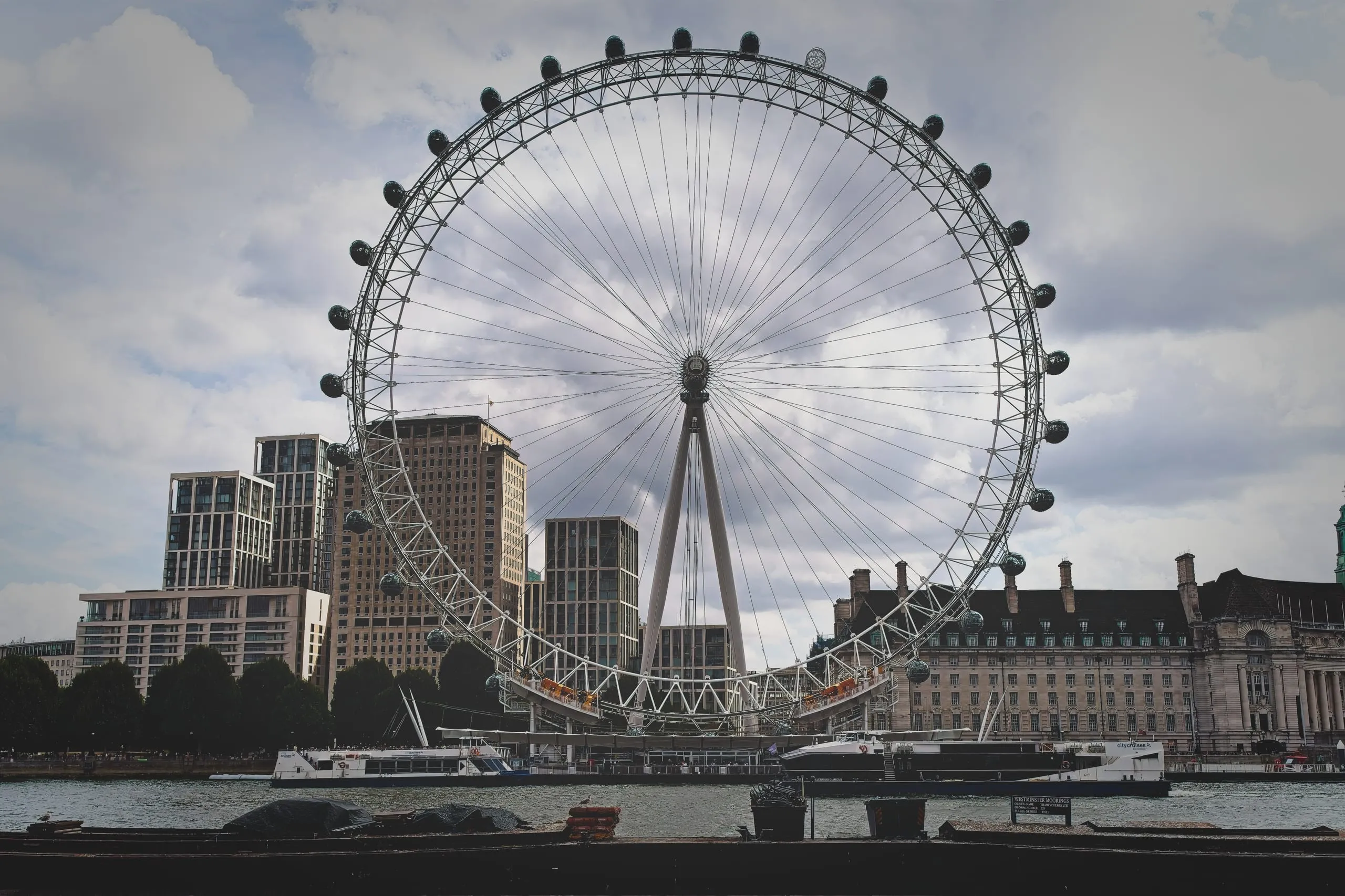Color photograph of the London Eye ferris wheel rising above the River Thames waterfront, with moored cruise boats and city towers in the background.