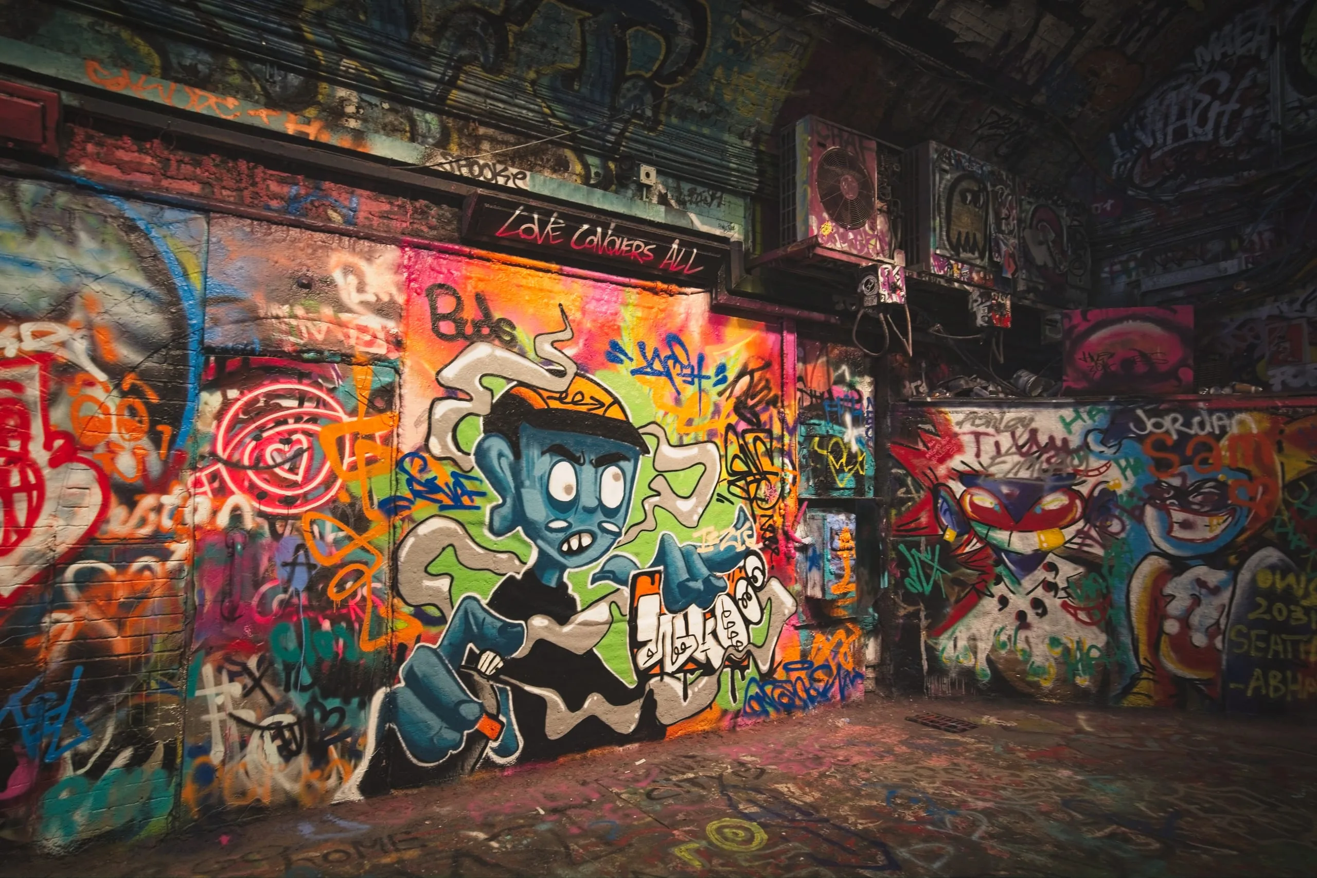 Photograph of a graffiti mural in London depicting a snarling blue cartoon character in a baseball cap, painted beneath a sign reading 'Love Conquers All' amid dense surrounding tags.