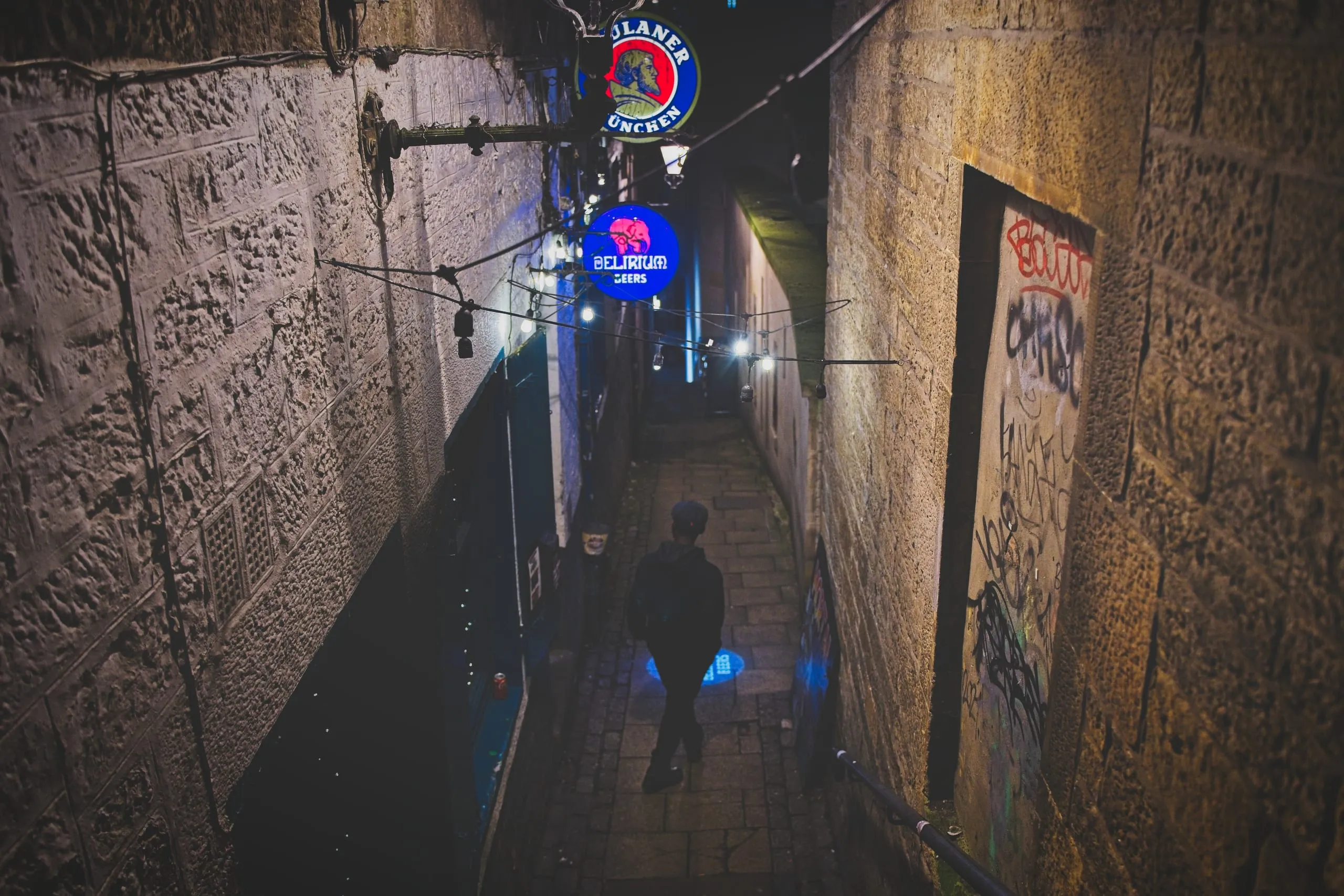 A color photograph of a narrow stone close in Edinburgh's Old Town at night, a lone figure walking beneath glowing neon pub signs and strings of bulb lights between graffiti-marked walls.