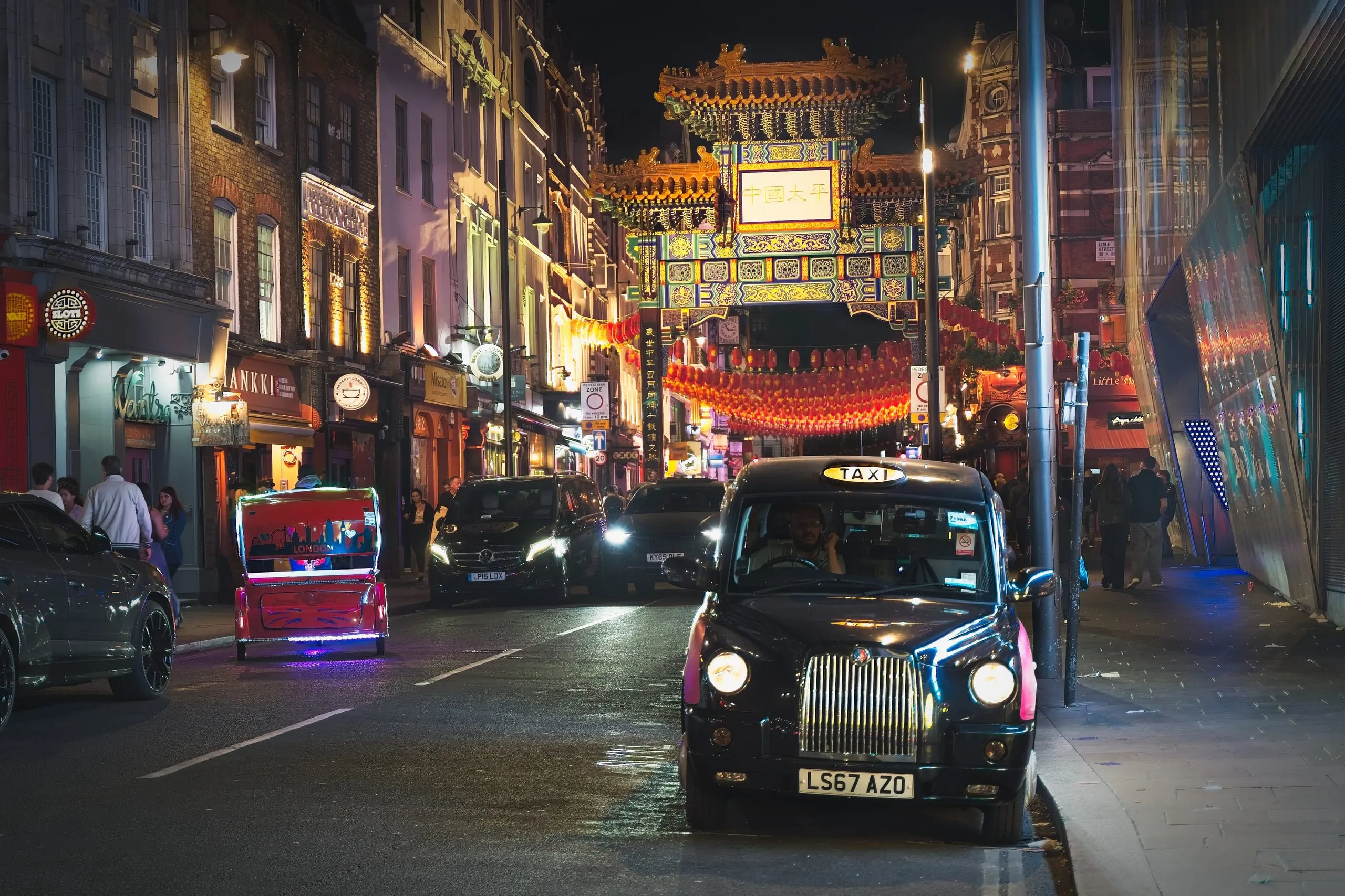 A night photograph of a black London taxi driving toward the illuminated Chinatown gate strung with red lanterns in London's West End.