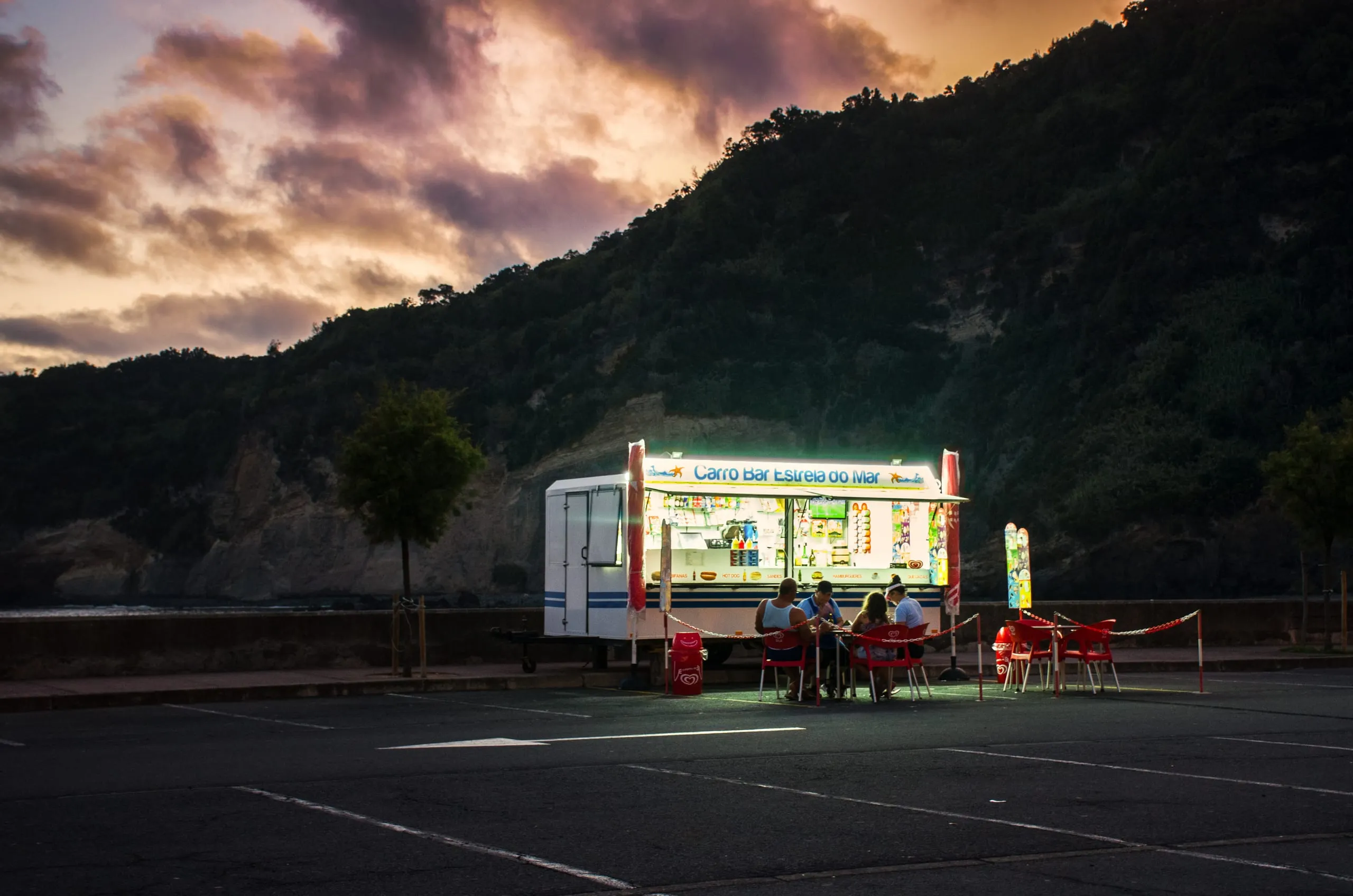 A brightly lit food-truck kiosk with red plastic chairs where a few people sit at dusk in an empty seafront parking lot below a dark ridge.