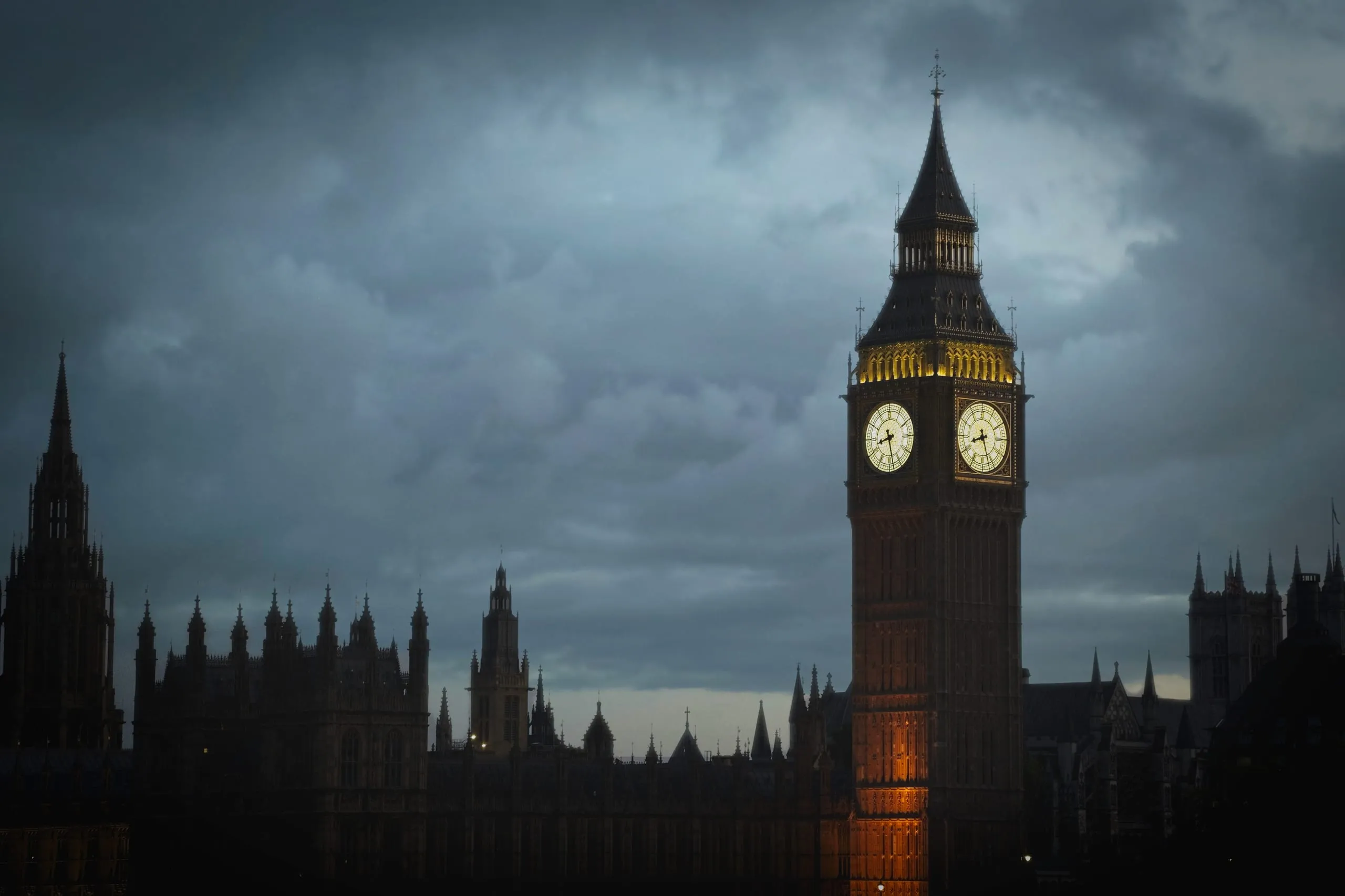 Photograph of Big Ben's illuminated clock tower glowing against a dark, storm-heavy evening sky above the silhouette of the Houses of Parliament, London, England.