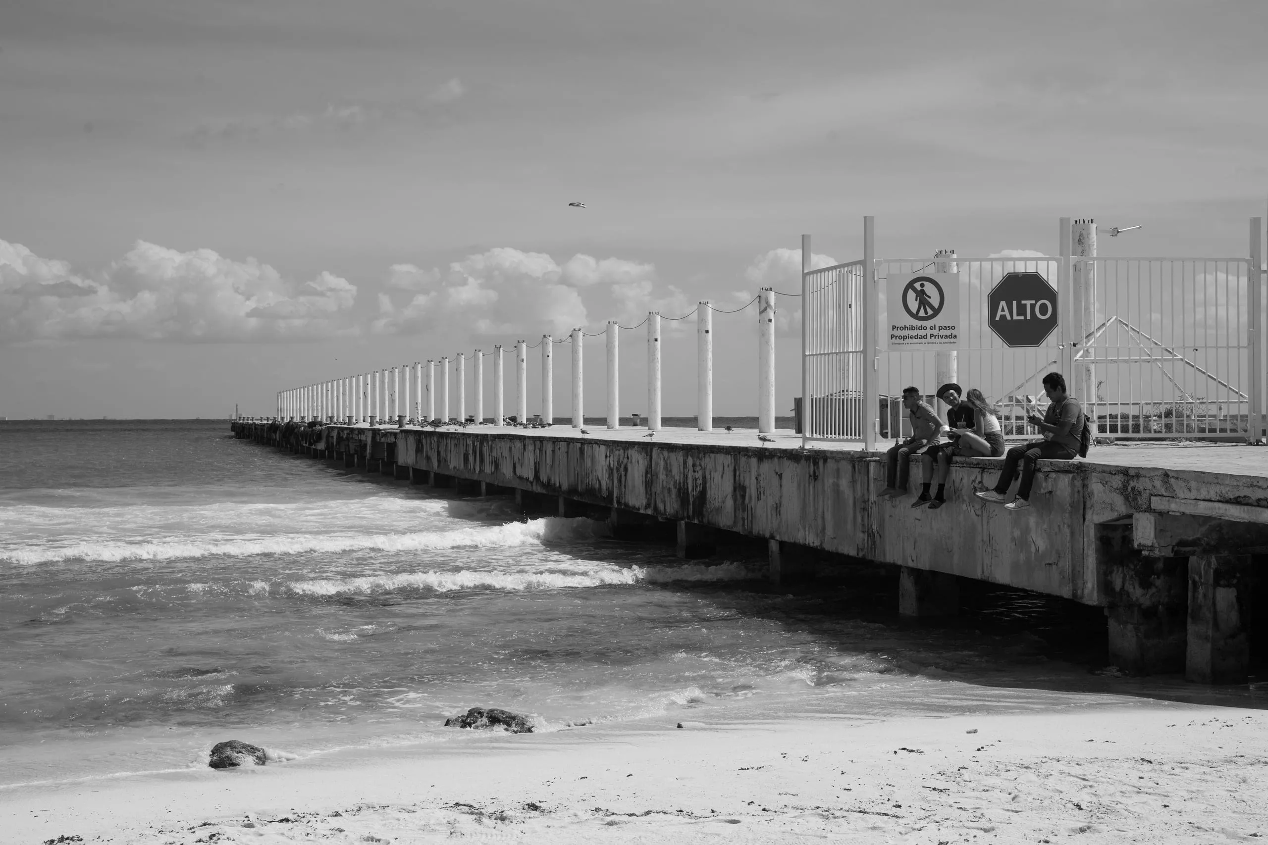 Black and white photograph of a long pier reaching into the Caribbean with people at its edge, Playa del Carmen