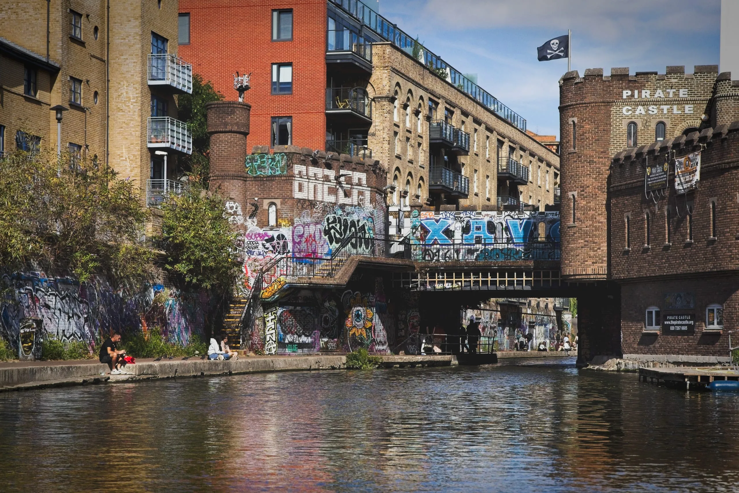 Color photograph of a graffiti-covered brick underpass on Regent's Canal in Camden, London, leading to the crenellated Pirate Castle building flying a skull-and-crossbones flag.