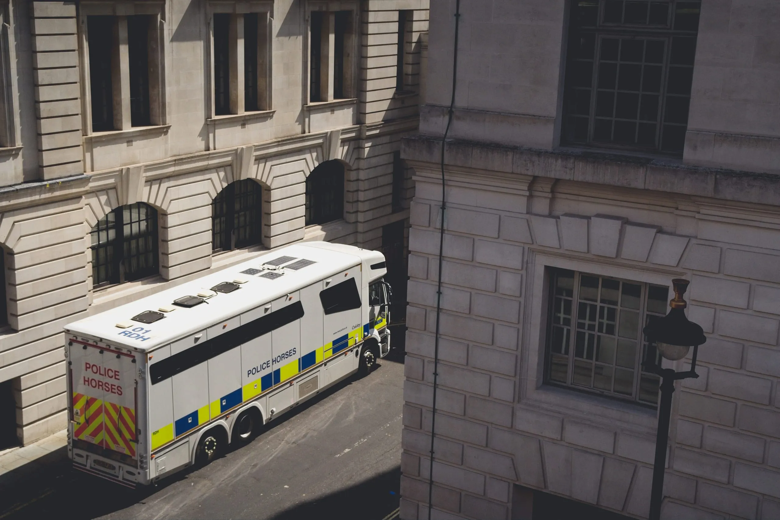 An elevated view of a white 'Police Horses' transport truck driving down a narrow street between grand stone government buildings in London, England.
