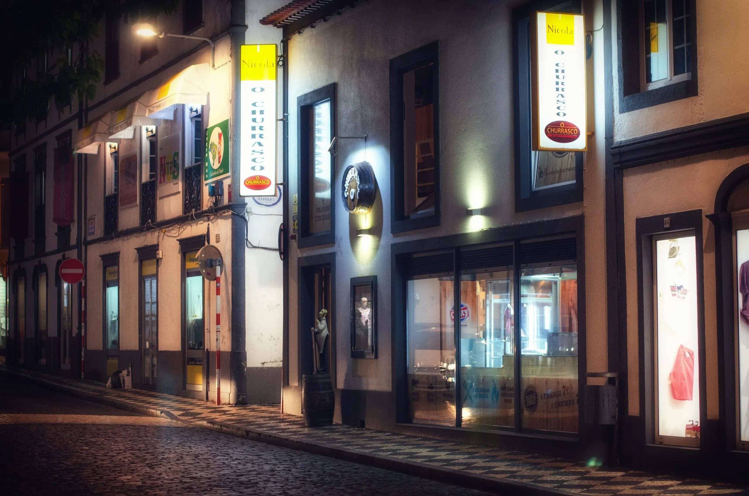 A narrow cobbled street at night with lit shop signs, a no-entry sign, and glowing storefront windows along old plastered buildings.