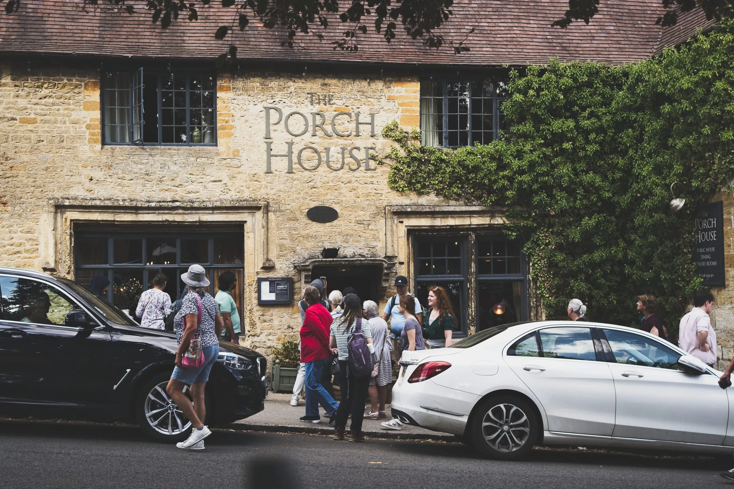 A photograph of tourists gathered outside The Porch House, a stone pub in Stow-on-the-Wold, the Cotswolds, England, with ivy on its facade and cars parked along the street.