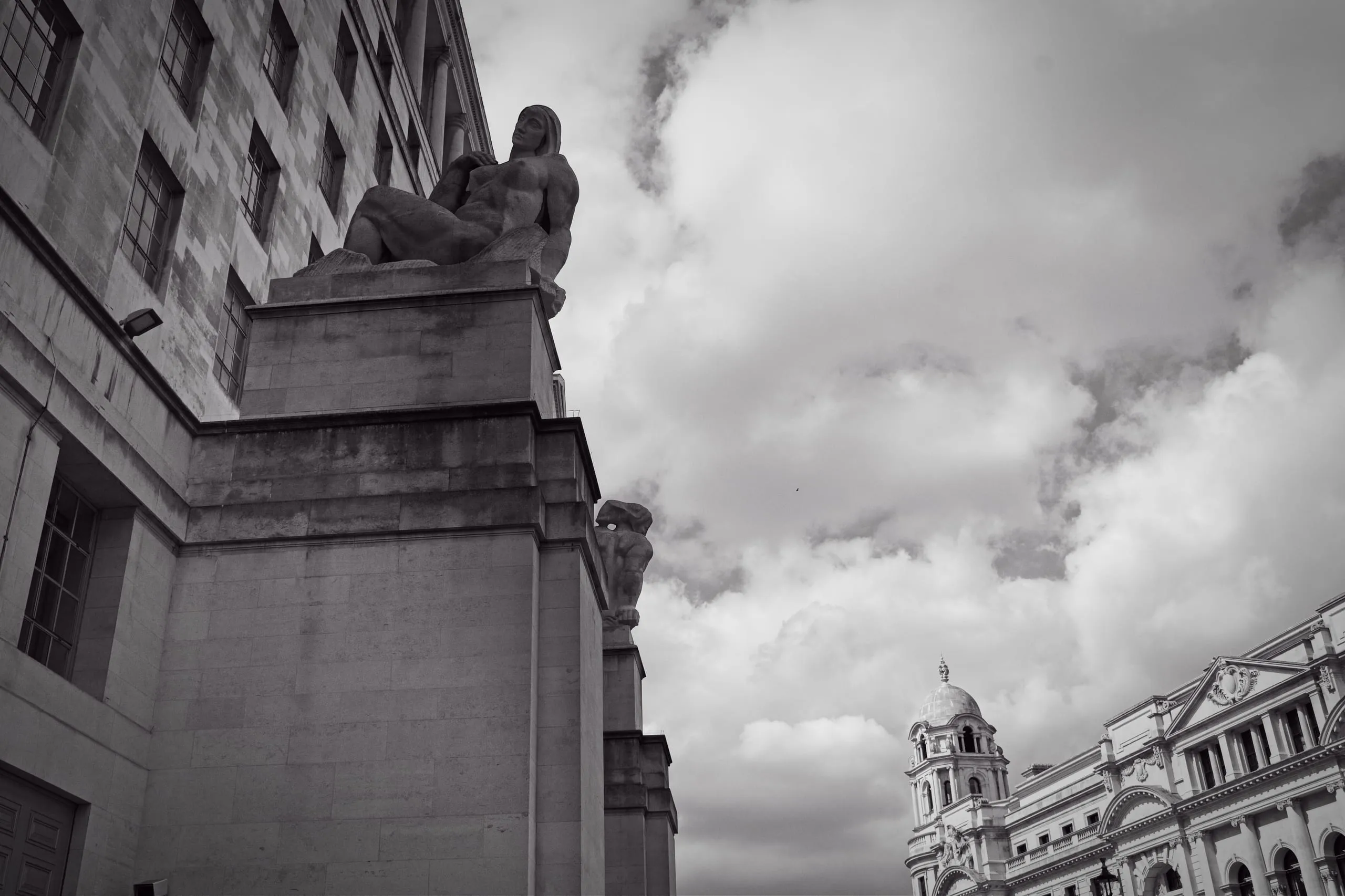 Black and white photograph of a monumental reclining stone figure carved into the corner of a London government building, set against a dramatic cloudy sky with a domed tower in the distance.