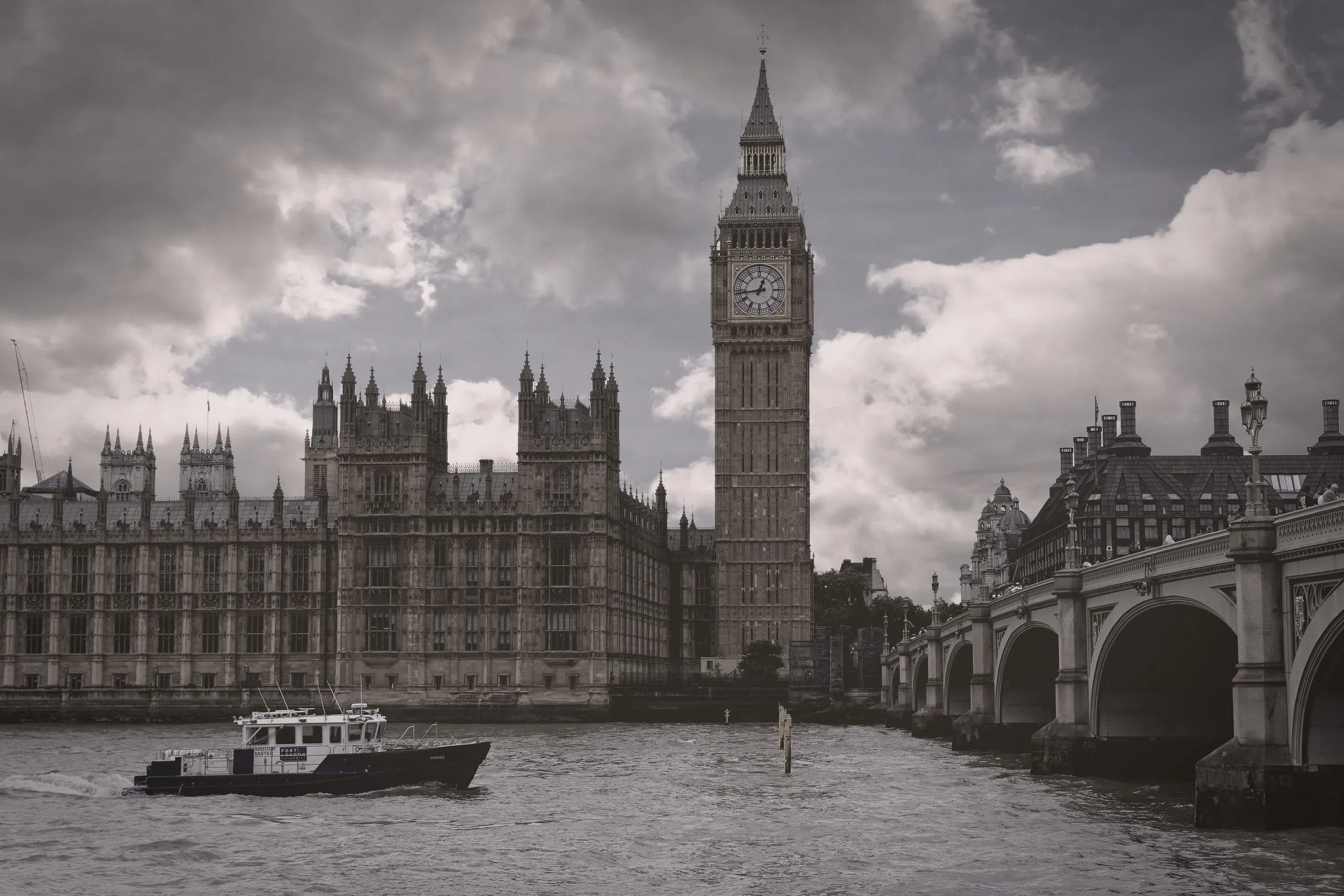 A harbor master's boat crosses the River Thames in front of the Houses of Parliament, Big Ben, and Westminster Bridge under a heavy, cloudy sky, London, England.