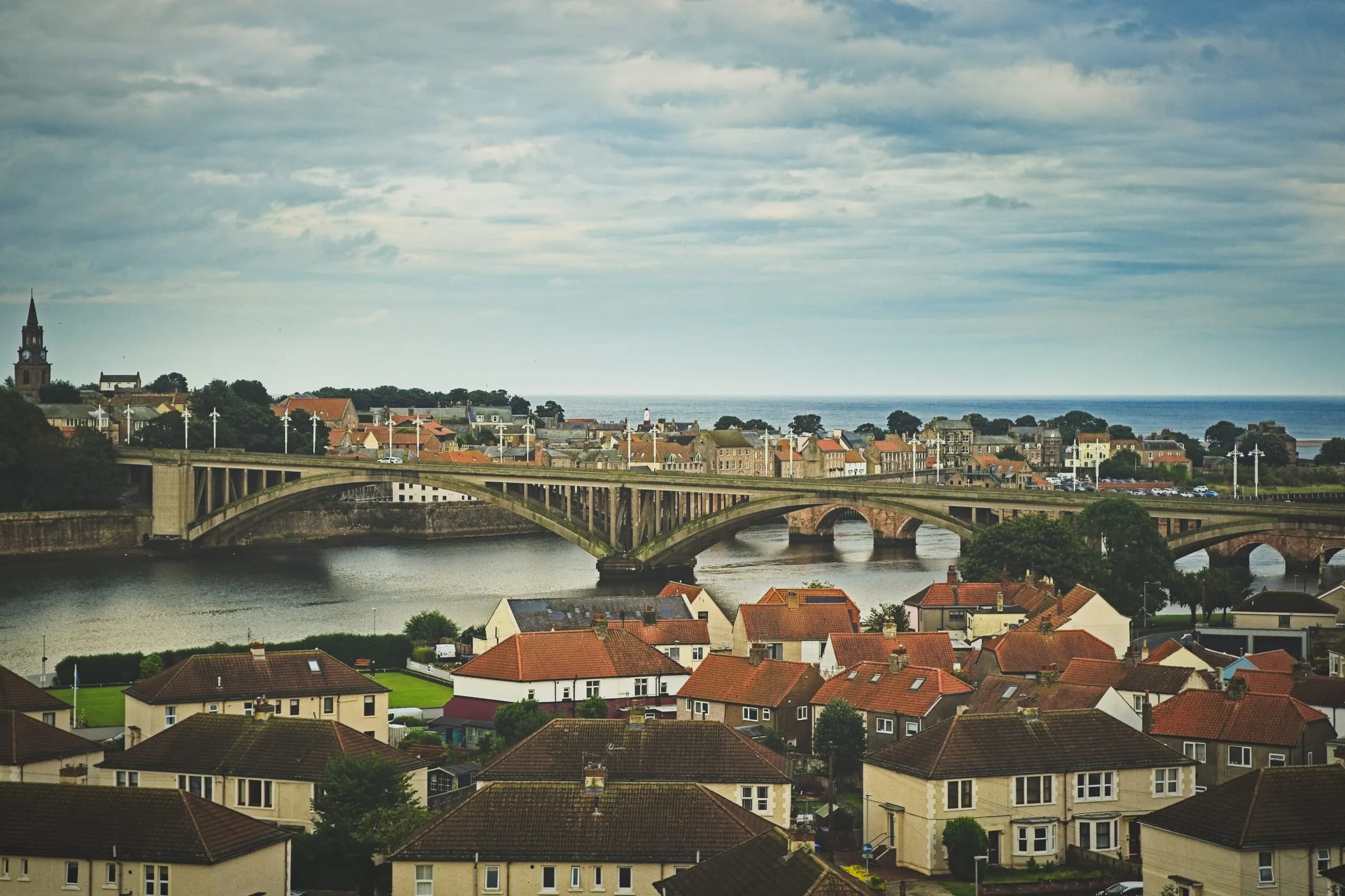 A color photograph of a river town in the Scottish Borders, with red-tiled rooftops in the foreground and an arched road bridge crossing the river toward the sea.