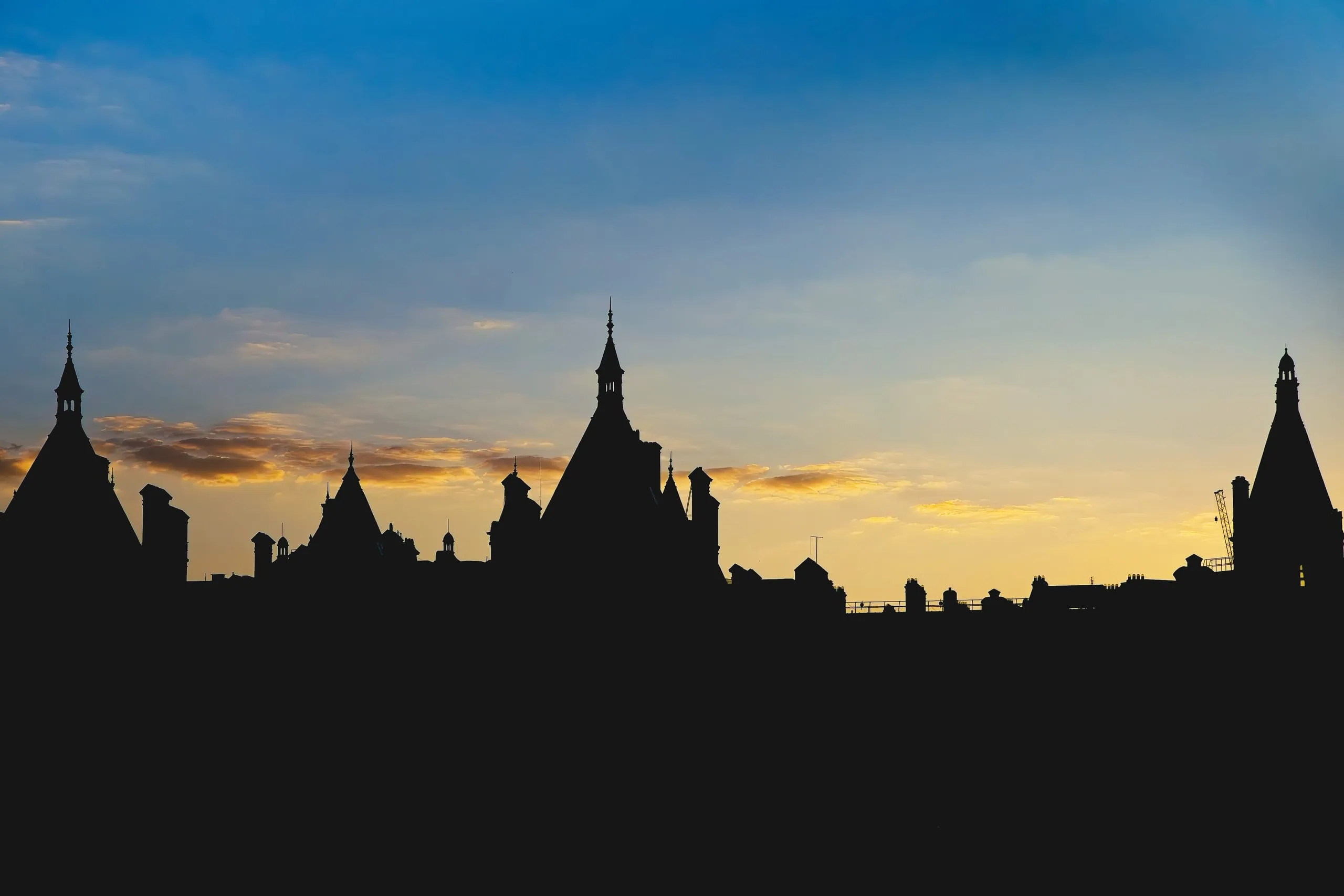 Photograph of the pointed turret rooftops of a Victorian building silhouetted against an orange and blue sunset sky, London, England.