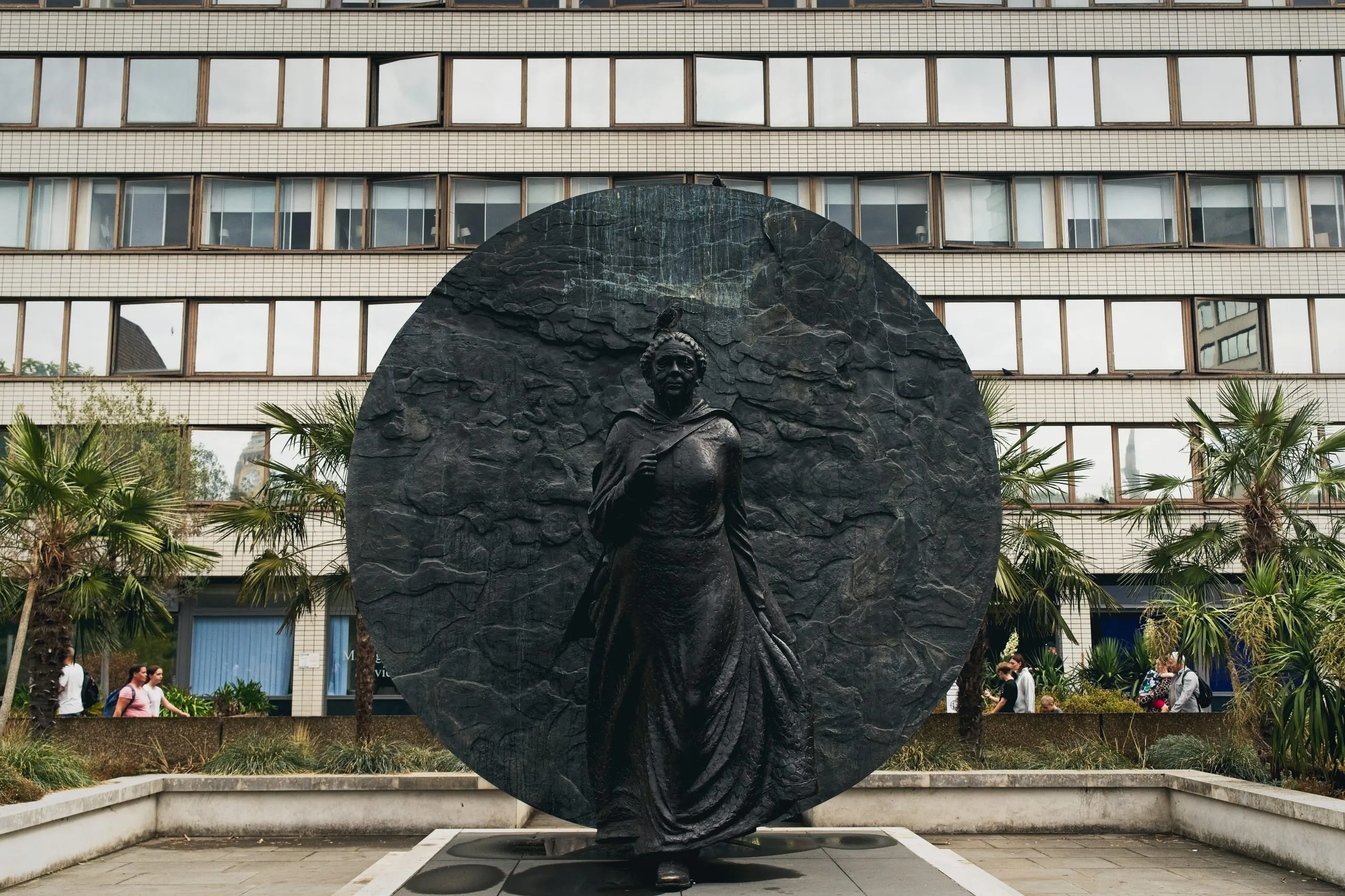 The Mary Seacole Memorial statue, a bronze figure of a woman in a cloak set against a large textured disc, flanked by palm trees in front of an office building near St Thomas' Hospital, London, England.