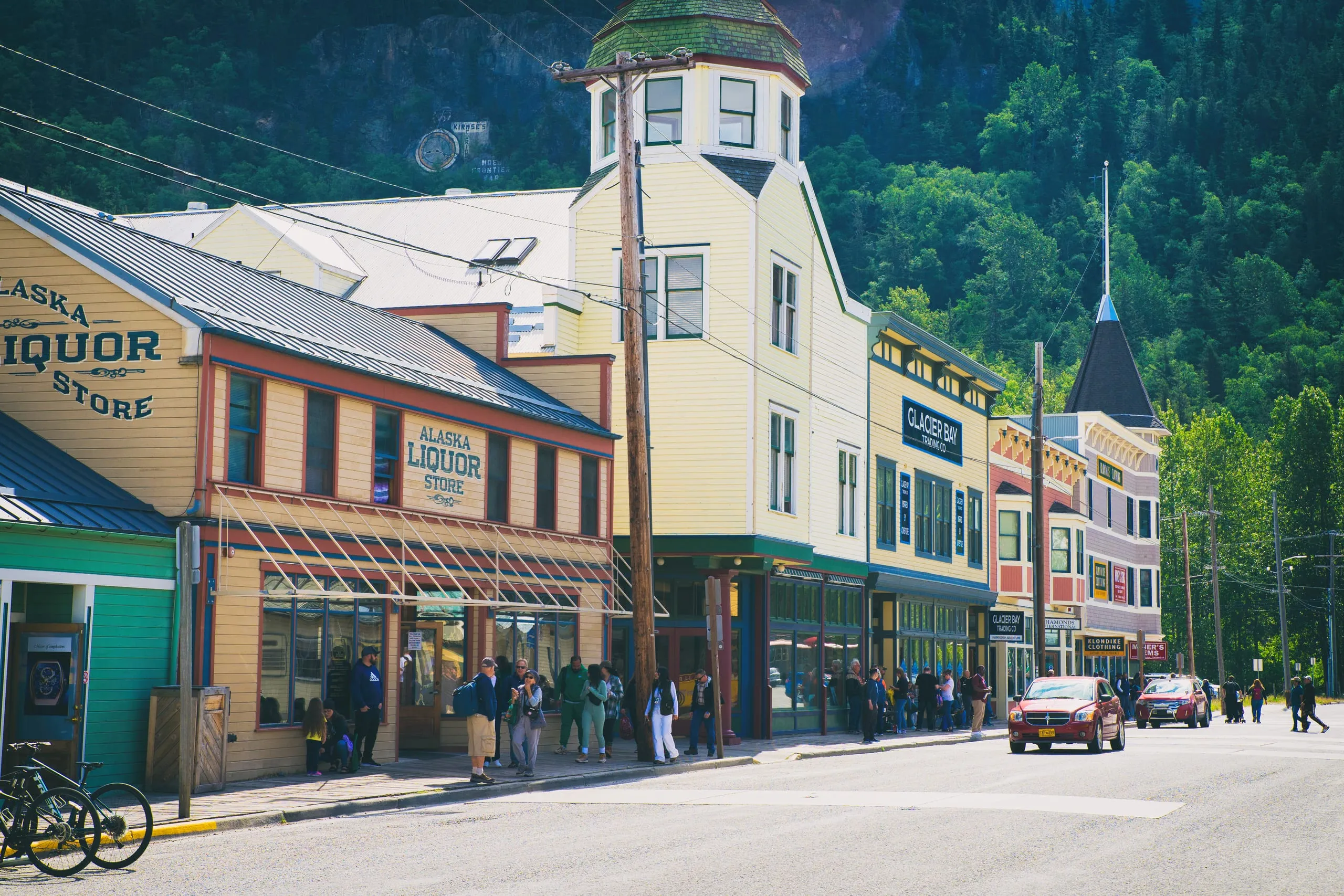 A sunlit block of restored gold-rush-era wooden storefronts with a corner turret building, people on the boardwalk and a red car passing.