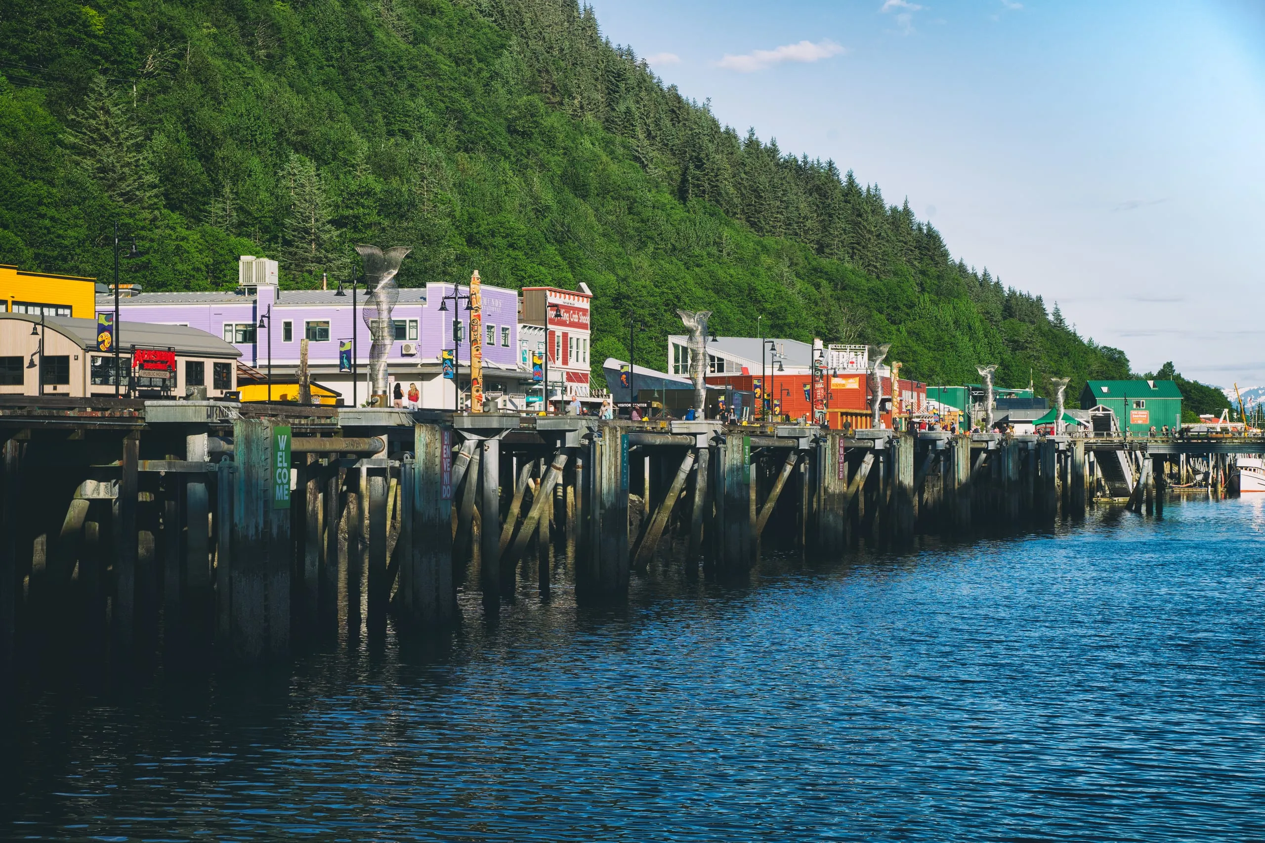 A row of colorful waterfront shops on a piling dock topped with silver whale-tail sculptures, backed by a steep spruce-covered hillside.