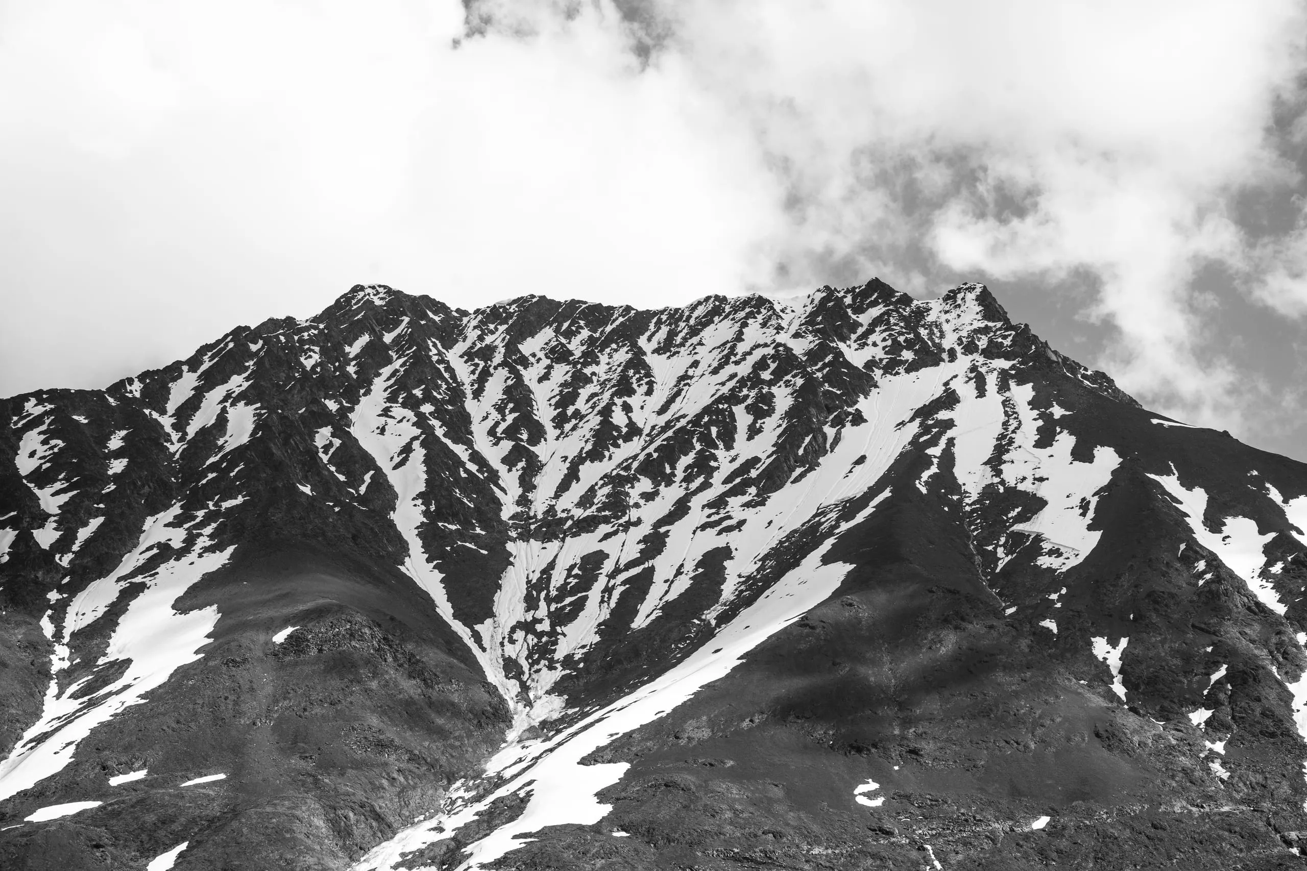 A black-and-white photograph of a rugged mountain peak streaked with patchy snow, its ridgeline dissolving into cloud.