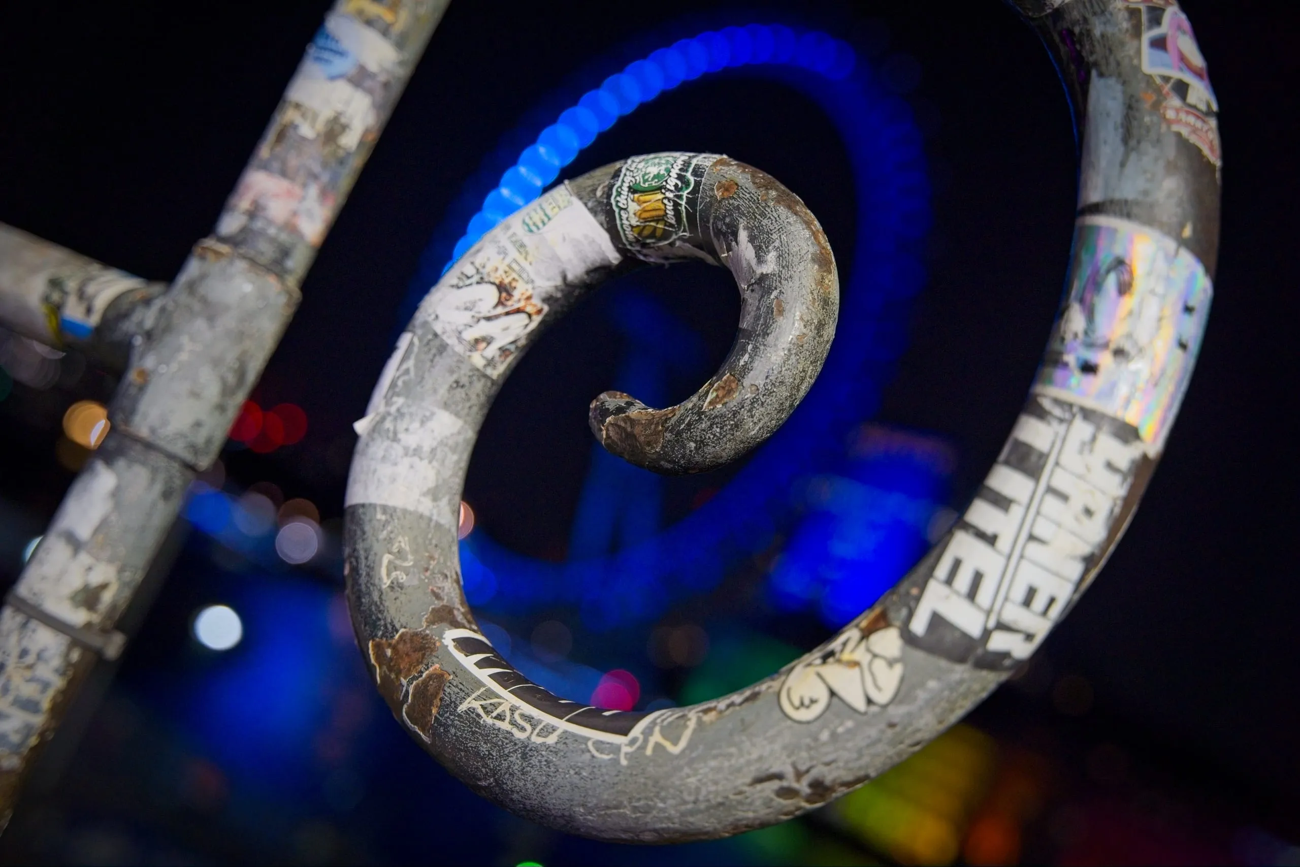 A photograph of a rusted, sticker-covered spiral metal railing at night, lit from behind by blue neon light in Camden, London, England.