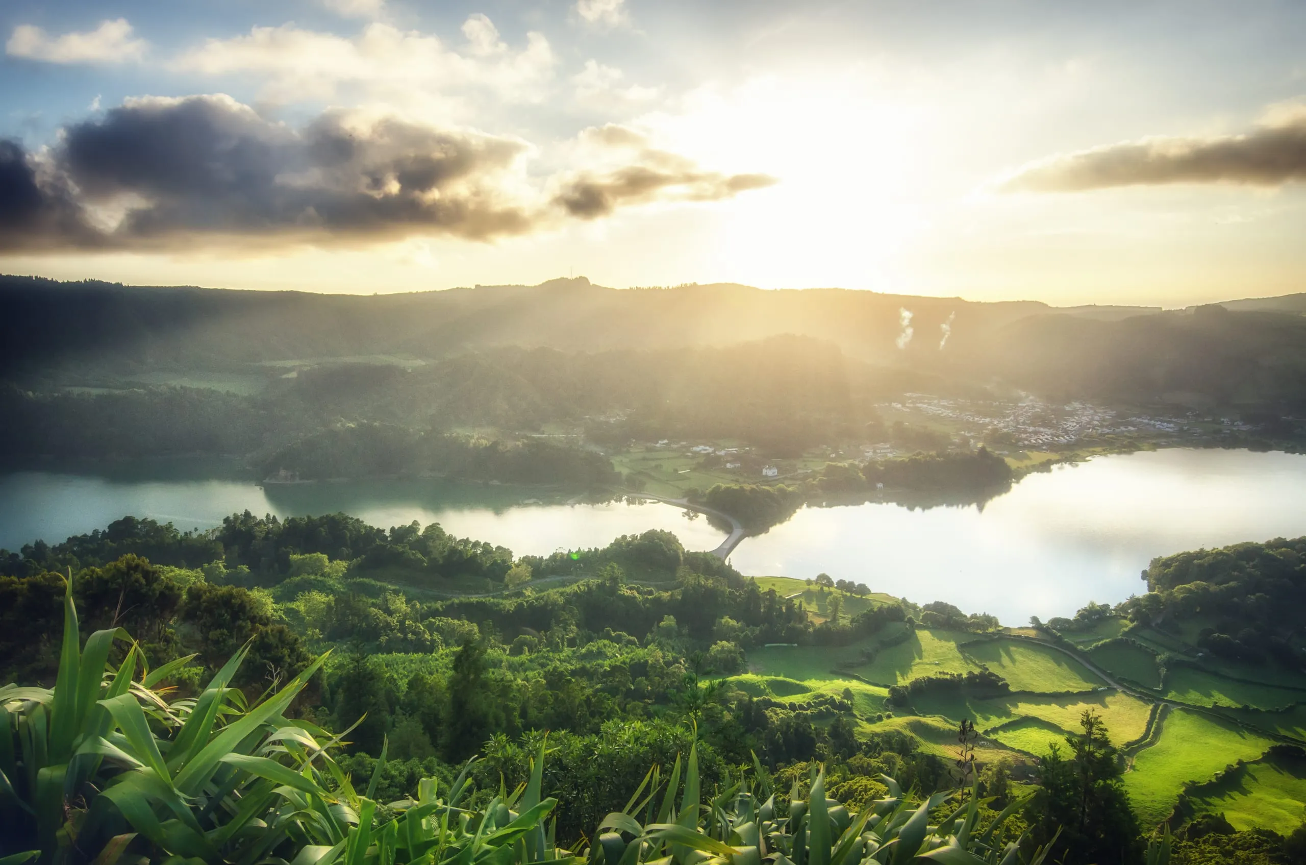 Sunlight breaks over ridges above twin crater lakes joined by a causeway, with a village and green fields below.