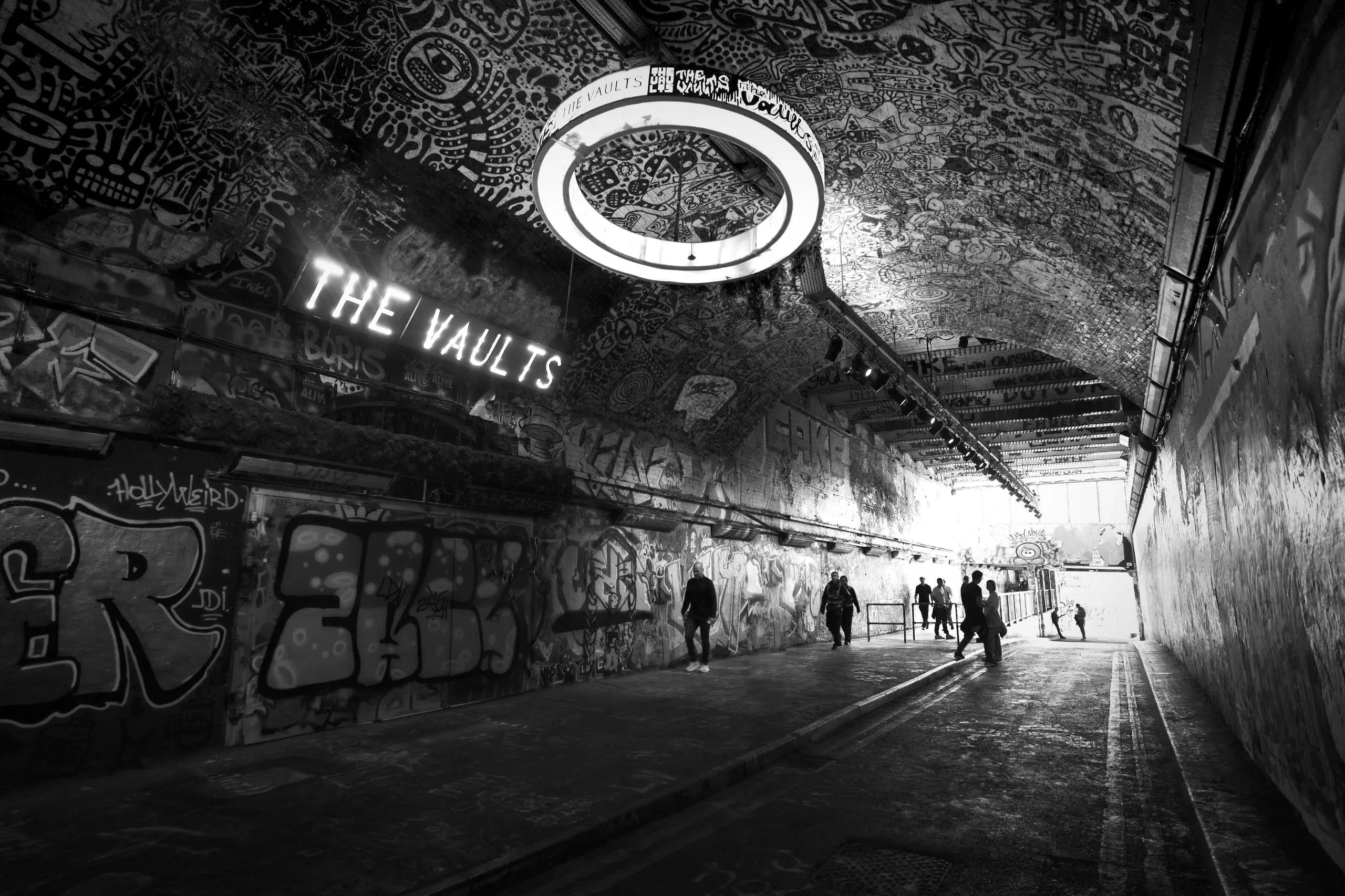 Black and white photograph of a graffiti-covered tunnel in London with an illuminated sign reading 'The Vaults' and a circular light fixture overhead, several pedestrians walking through.